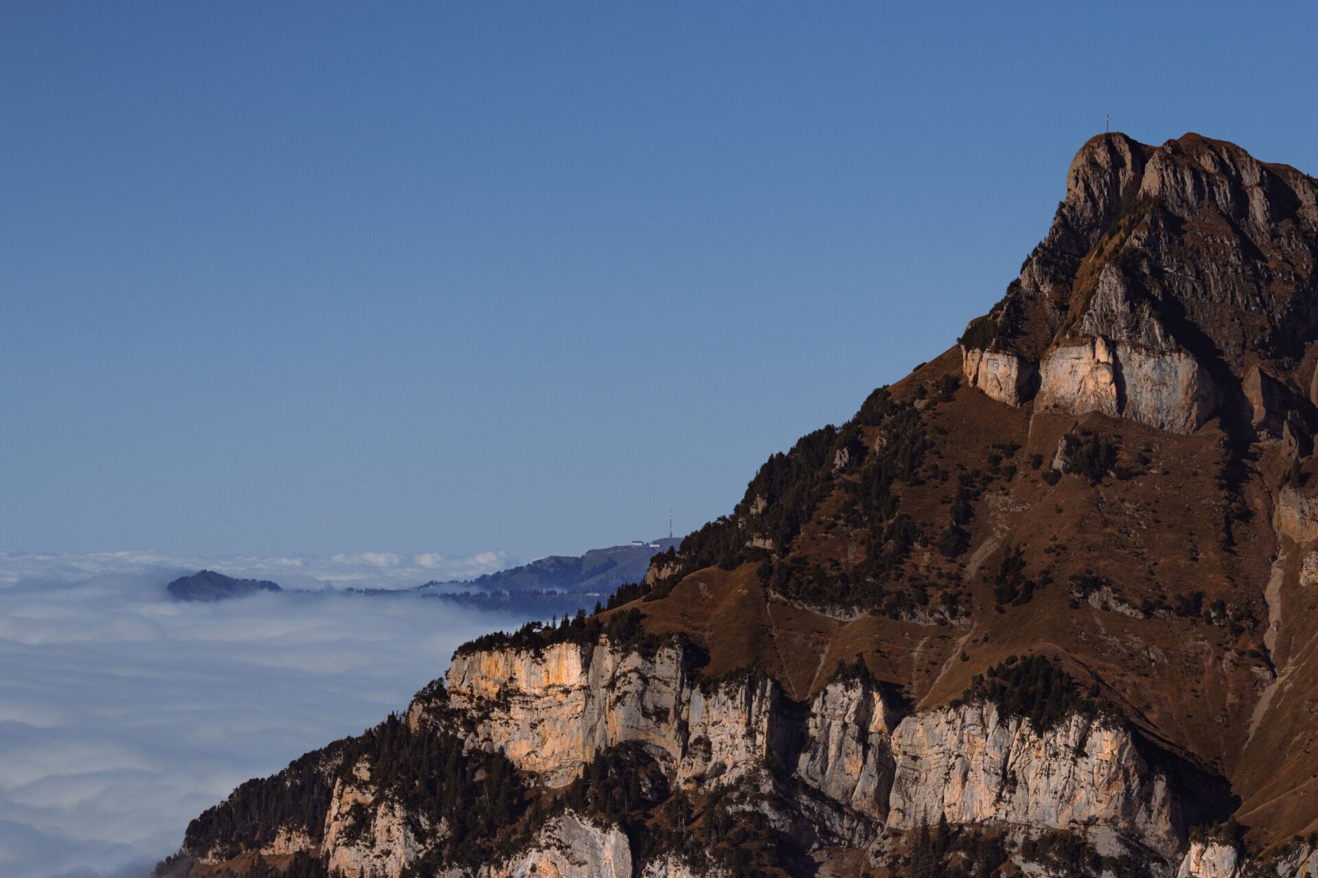 Ein markanter, felsiger Berg mit einem Gipfelkreuz ragt über ein weites Wolkenmeer, das wie ein ruhiger, weißer Ozean die tiefer liegenden Täler bedeckt. Die warmen Farbtöne des Gesteins kontrastieren eindrucksvoll mit dem klaren Blau des Himmels und verleihen der Szene eine majestätische Ruhe.