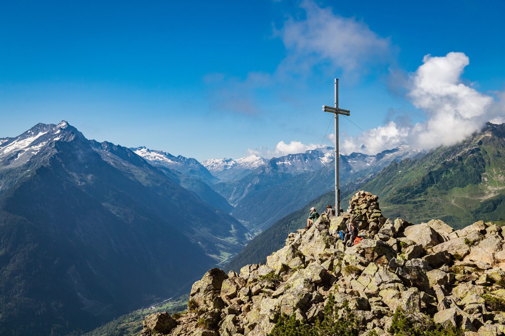 Ein Gipfelkreuz thront auf einer felsigen Anhöhe, umgeben von beeindruckenden, schneebedeckten Bergen und einem tiefen Tal. Die klare Luft und das majestätische Panorama schaffen eine erhabene und friedliche Atmosphäre.