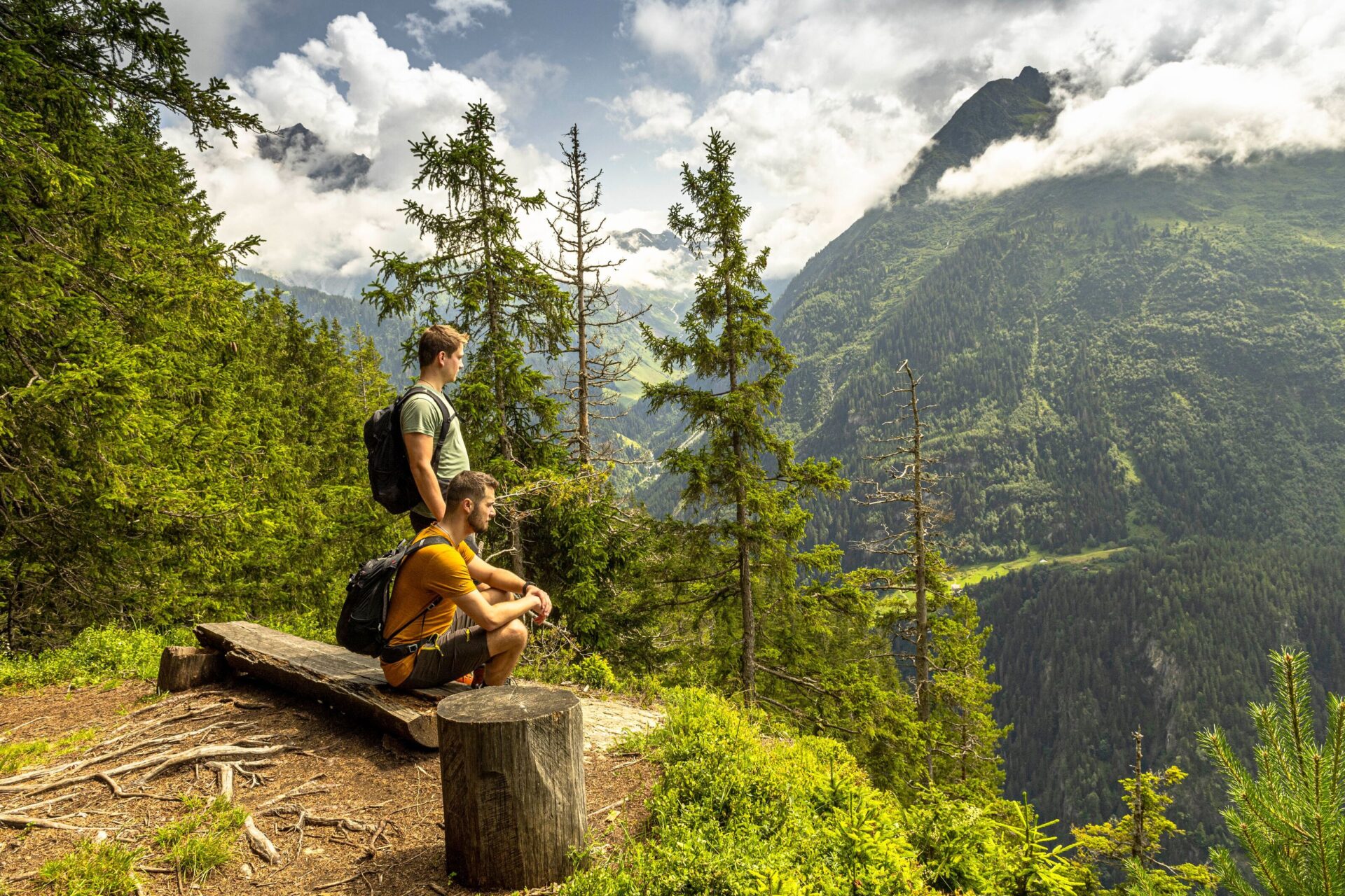 Zwei Wanderer machen auf dem Höhenweg in den Silenerbergen eine Pause auf einer einfachen Holzbank und blicken über bewaldete Hänge in ein weites Bergtal. Die erhöhte Lage, das weiche Licht und die offene Aussicht vermitteln Ruhe, Weite und ein intensives Naturerlebnis.