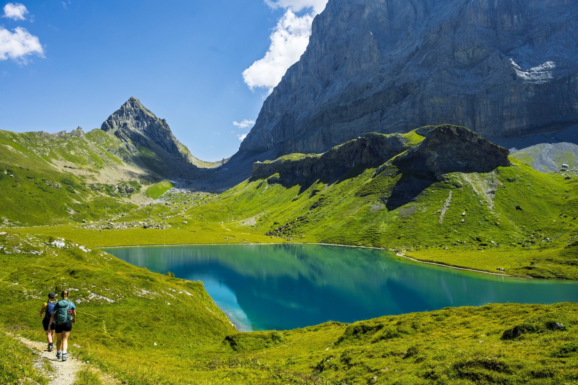 Ein türkisblauer Bergsee liegt eingebettet zwischen saftig grünen Hängen und imposanten Felswänden unter strahlend blauem Himmel. Zwei Wandernde folgen einem schmalen Pfad entlang der idyllischen Hochgebirgslandschaft und verleihen der Szene eine ruhige, naturnahe Stimmung.