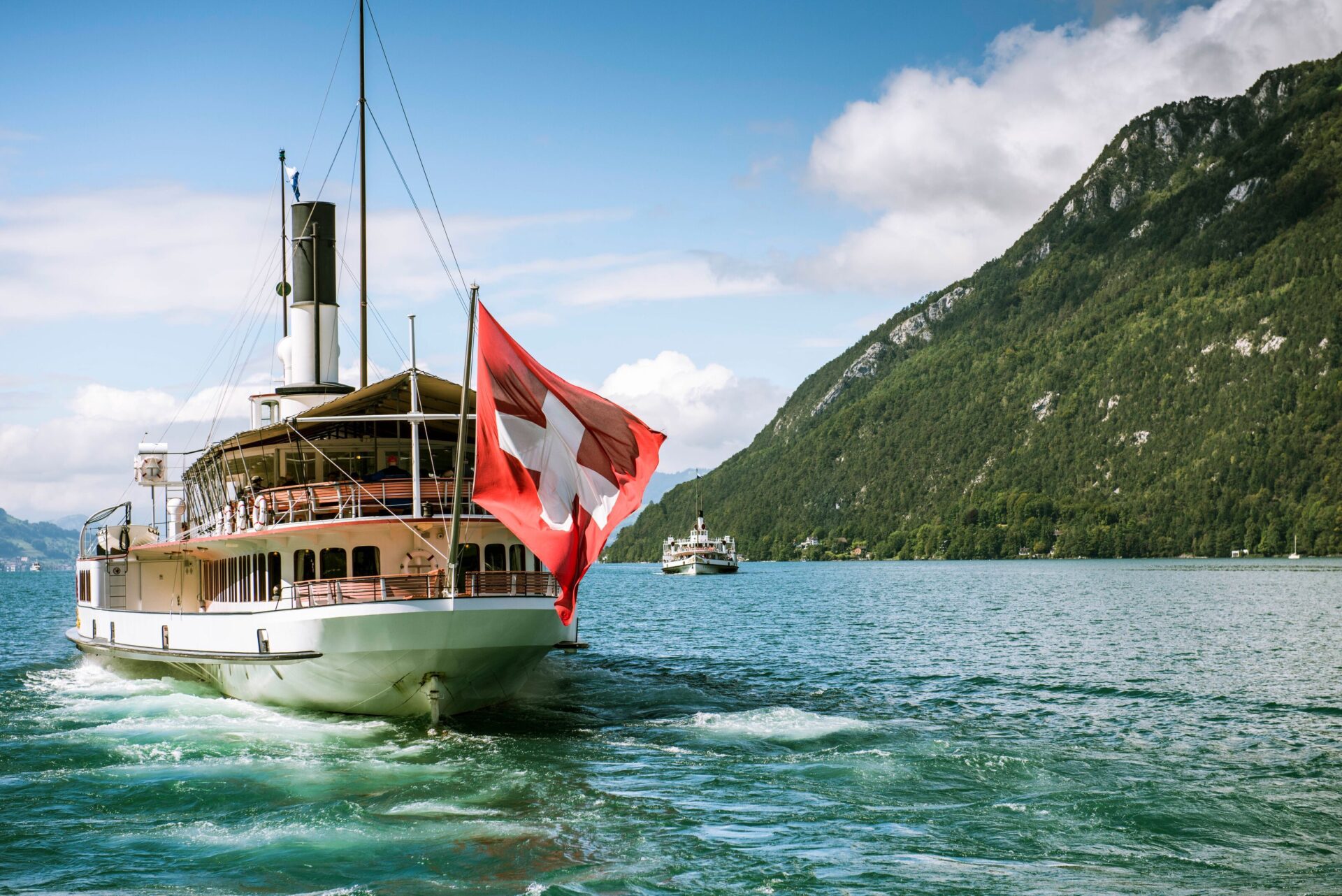 Ein traditionelles Dampfschiff mit einer großen Schweizer Flagge am Heck fährt über einen klaren, ruhigen See, umgeben von grünen Bergen und bewölktem Himmel. Die Szenerie strahlt Ruhe und Naturverbundenheit aus, während das Schiff friedlich auf dem Wasser gleitet.