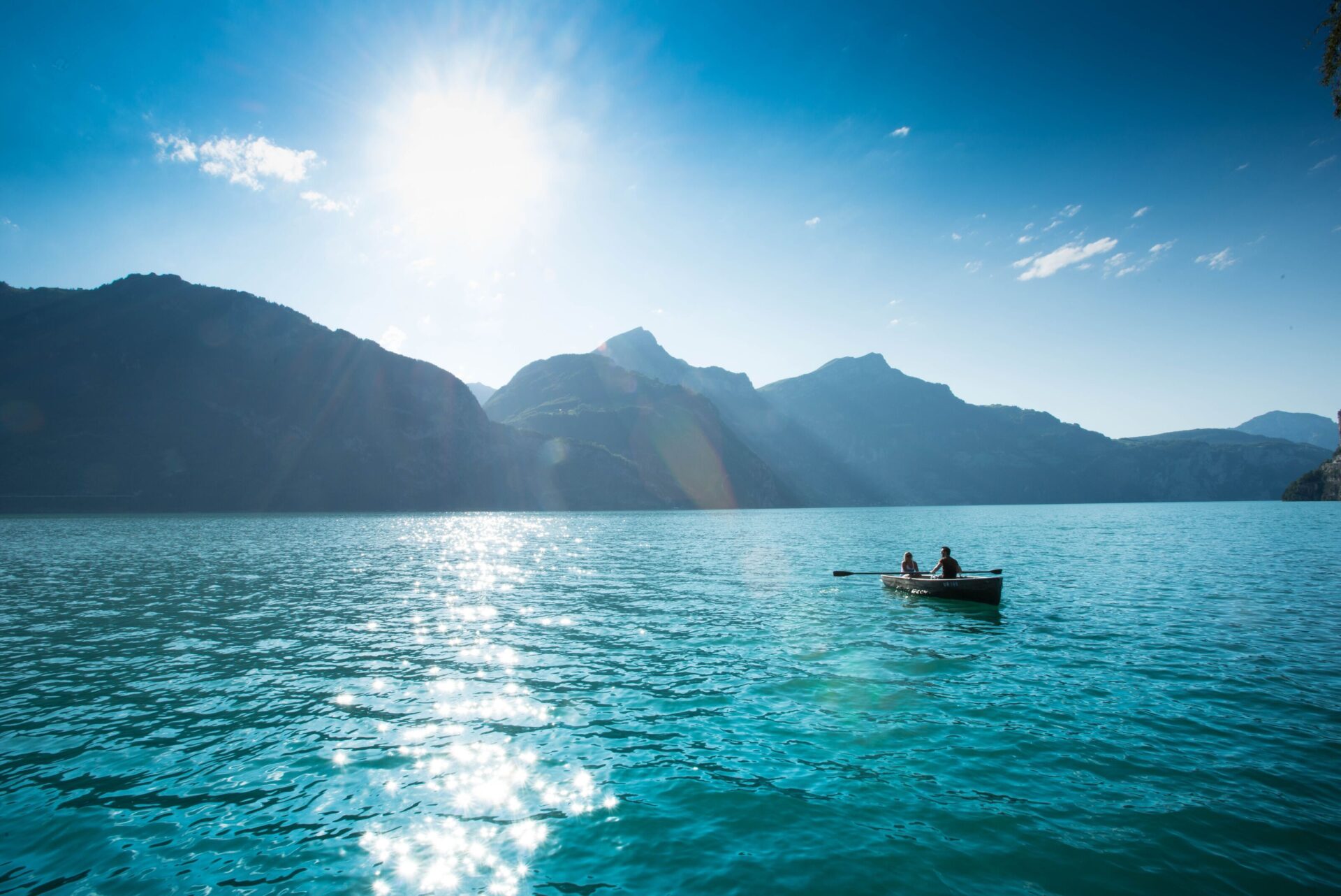 Zwei Menschen rudern in einem kleinen Boot auf einem türkisblauen See, während die Sonne hoch am Himmel steht und das Wasser glitzern lässt. Die umgebenden Berge und das strahlende Licht schaffen eine friedliche und idyllische Szenerie.