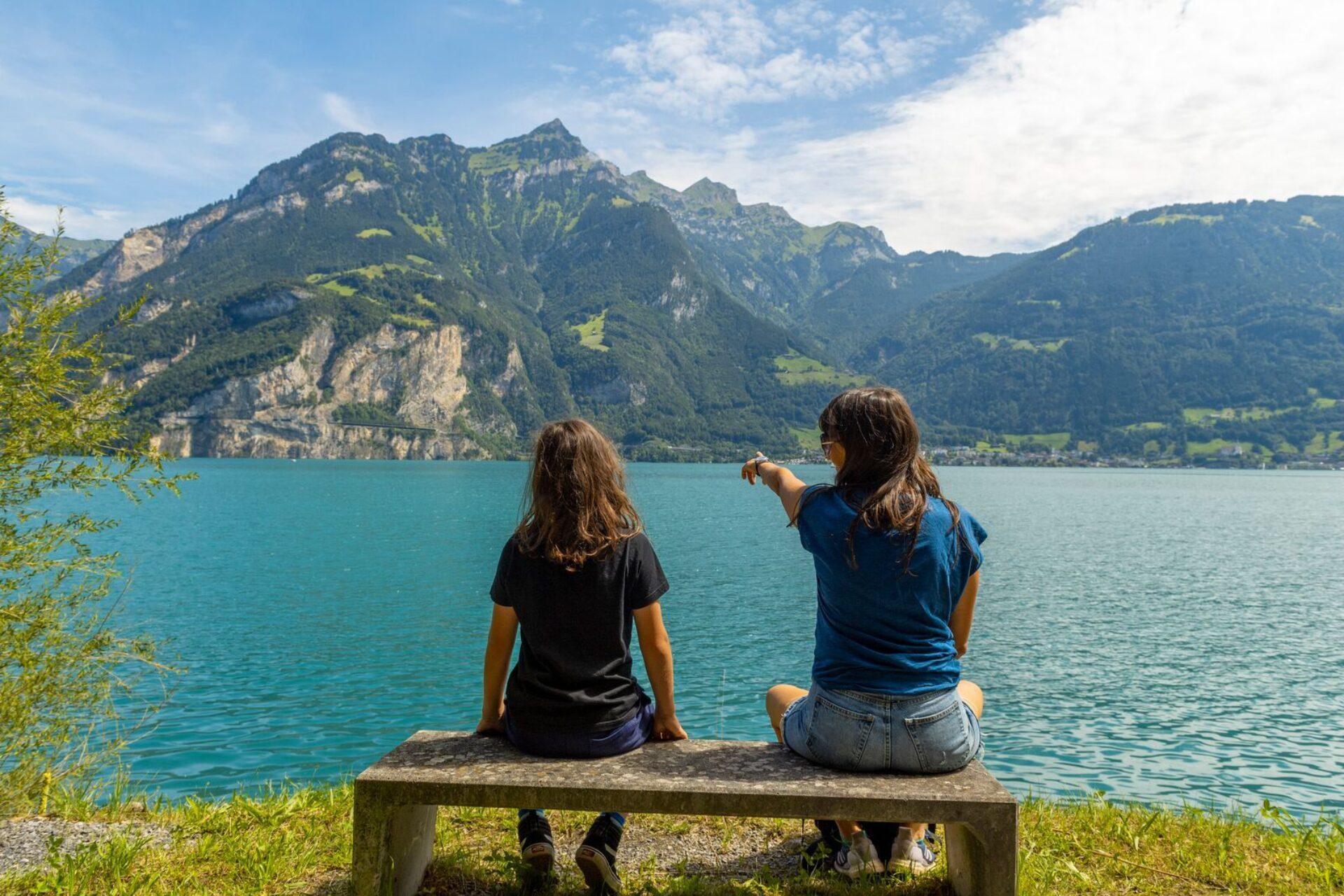 Zwei Personen sitzen auf einer Holzbank am Ufer eines türkisblauen Sees und blicken auf die majestätischen, grünen Berge vor ihnen. Eine der beiden zeigt in die Ferne, während die friedliche Landschaft und das klare Wasser eine ruhige und entspannte Atmos