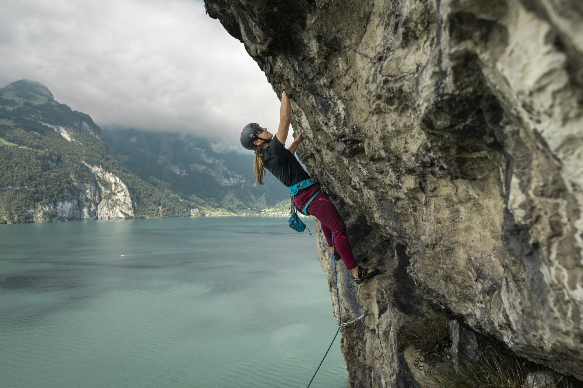 Eine Kletterin hängt konzentriert an einer steilen Felswand hoch über einem türkisfarbenen See, während im Hintergrund nebelverhangene Berge und dichte Wälder eine dramatische Kulisse bilden. Die Szene vermittelt Kraft, Mut und die Nähe zur unberührten Natur.