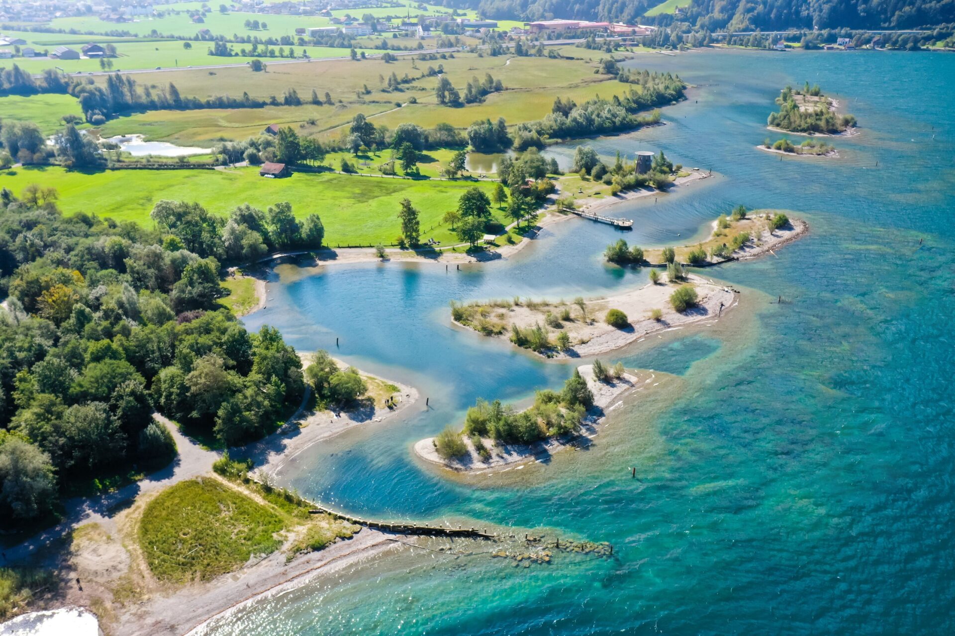 Eine malerische Landschaft aus der Vogelperspektive zeigt eine Küstenlinie mit kleinen Inseln und grün bewachsenen Landzungen, die ins türkisfarbene Wasser eines Sees ragen. Im Hintergrund erstrecken sich grüne Wiesen und Felder, die eine harmonische Verbindung zwischen Land und Wasser schaffen.