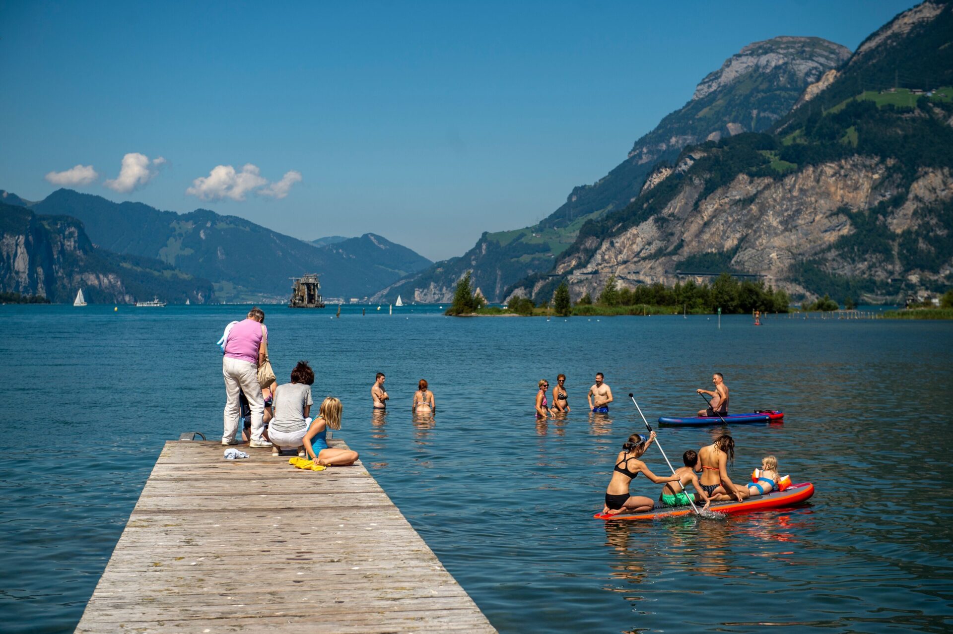 Menschen genießen einen sonnigen Tag an einem See, schwimmen im klaren Wasser und fahren auf Kajaks, während im Vordergrund ein Steg ins Wasser ragt. Umgeben von imposanten Bergen vermittelt die Szene pure Sommerfreude und Erholung in der Natur.