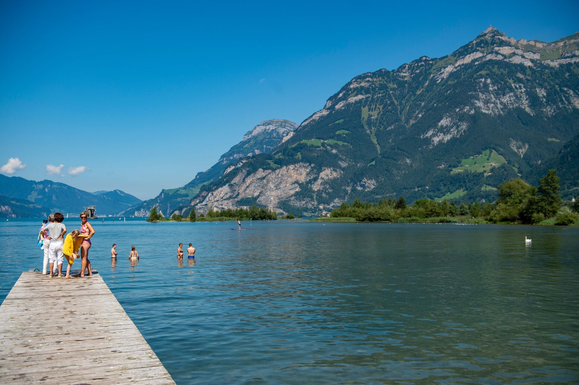 Mehrere Menschen genießen einen Sommertag am See, einige stehen auf einem Holzsteg, während andere im Wasser schwimmen. Die umliegenden Berge und der strahlend blaue Himmel schaffen eine idyllische Urlaubsatmosphäre.