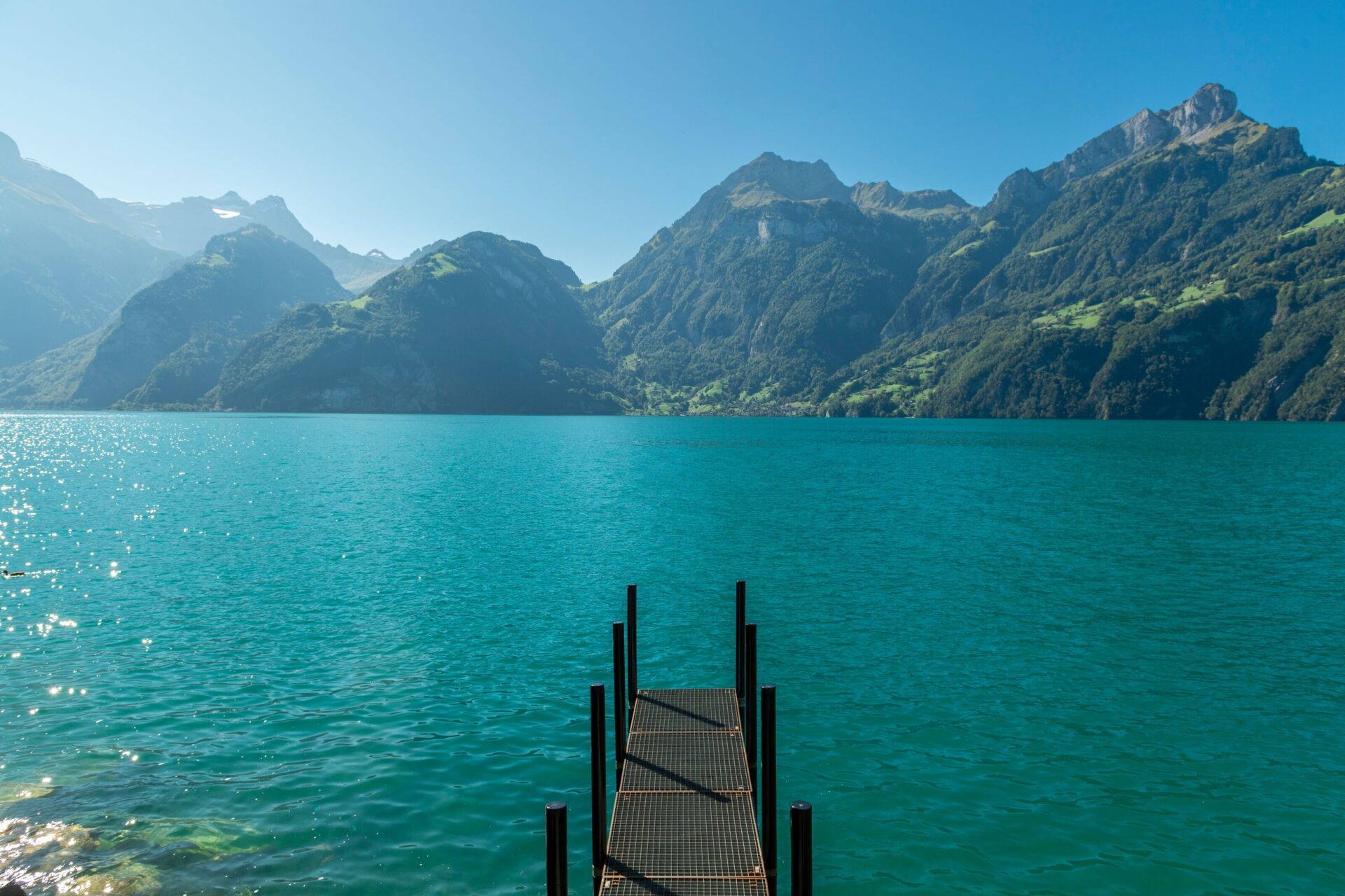 Ein kleiner Steg ragt in das klare, türkisfarbene Wasser eines Sees, umgeben von majestätischen Bergen unter einem strahlend blauen Himmel. Die Szenerie strahlt Ruhe und die beeindruckende Schönheit der Natur aus.