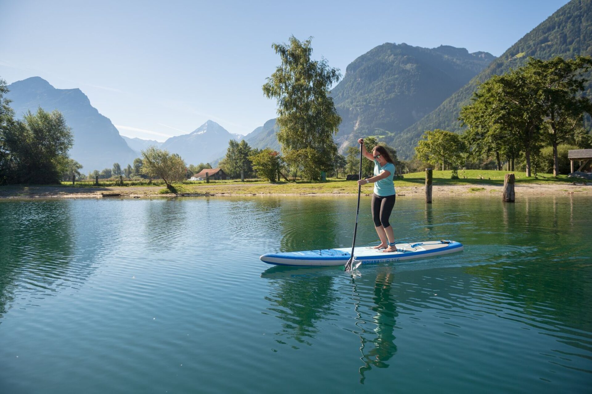Eine Frau paddelt entspannt auf einem ruhigen See, umgeben von grünen Bäumen und hohen Bergen im Hintergrund. Die friedliche Landschaft und das klare Wasser schaffen eine Atmosphäre von Ruhe und Naturverbundenheit.