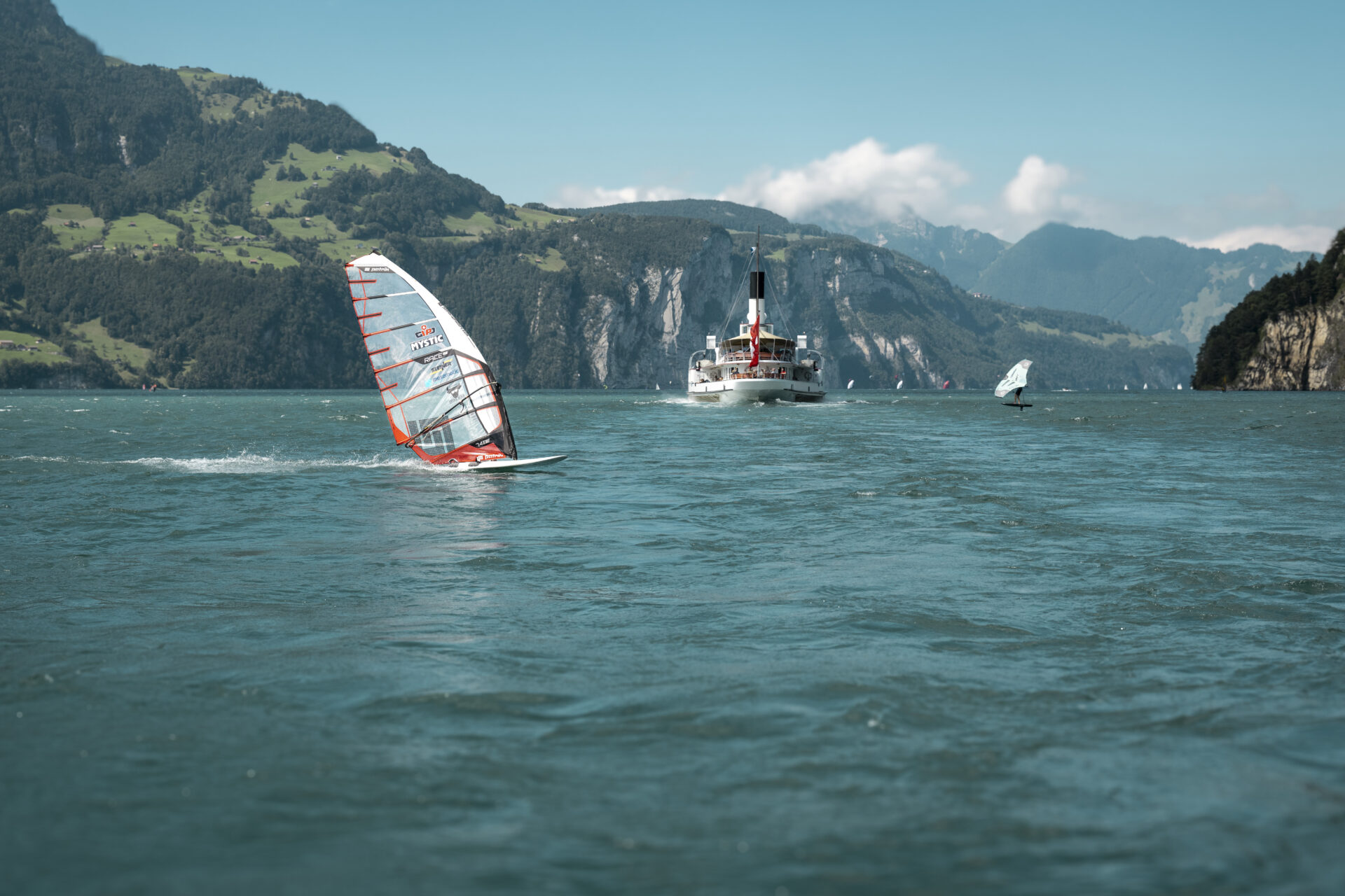 Ein einzelner Windsurfer zieht seine Bahn über einen ruhigen Bergsee, während sich im Hintergrund ein dramatisch beleuchtetes Alpenmassiv mit wolkenverhülltem Gipfel erhebt. Die Szenerie vermittelt ein Gefühl von Freiheit, Ruhe und der gewaltigen Schönheit der Natur.