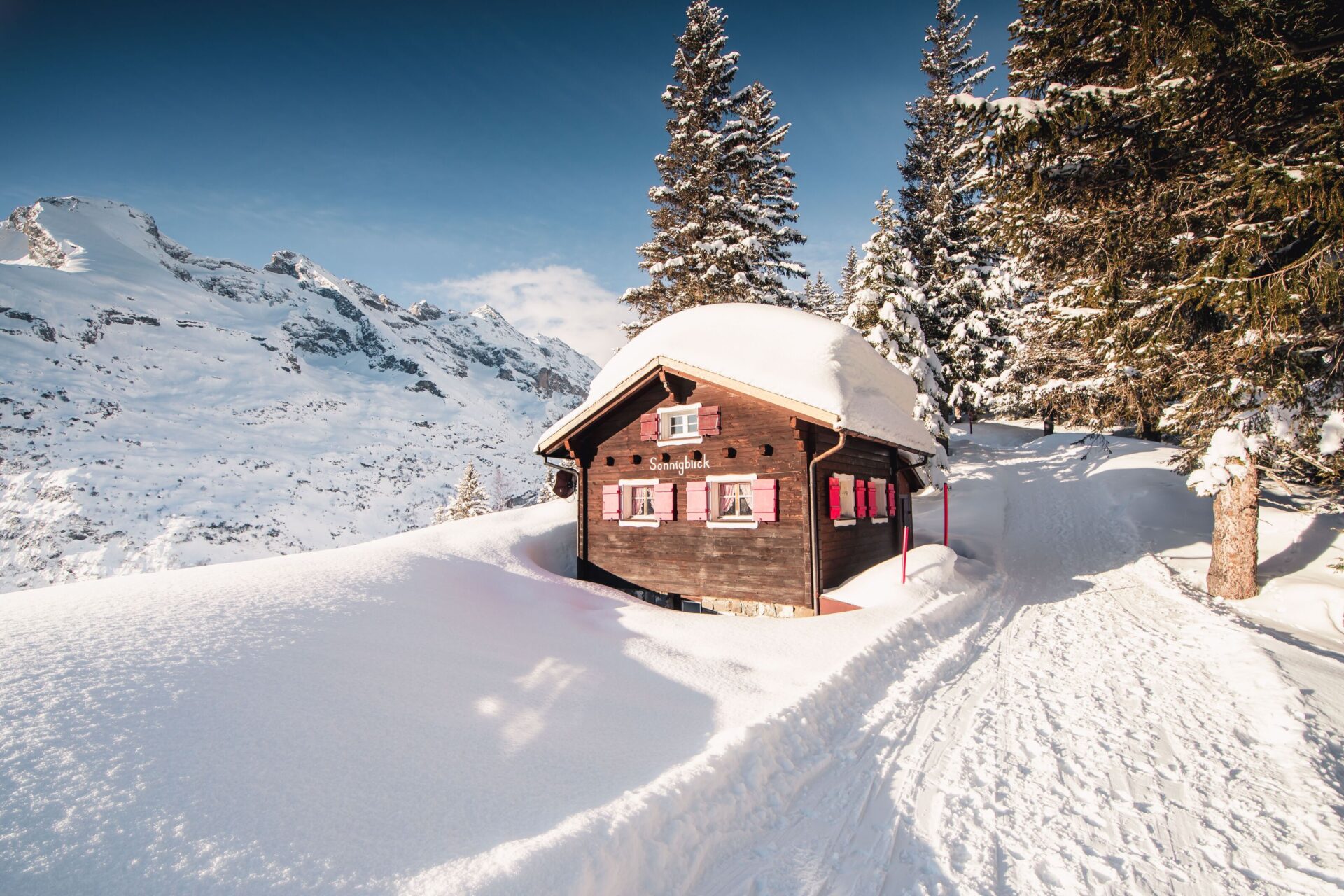 Eine malerische Berghütte mit schneebedecktem Dach steht idyllisch zwischen verschneiten Bäumen, umgeben von einer ruhigen Winterlandschaft und majestätischen Bergen. Die klare Luft und das sanfte Licht schaffen eine friedliche und einladende Atmosphäre.