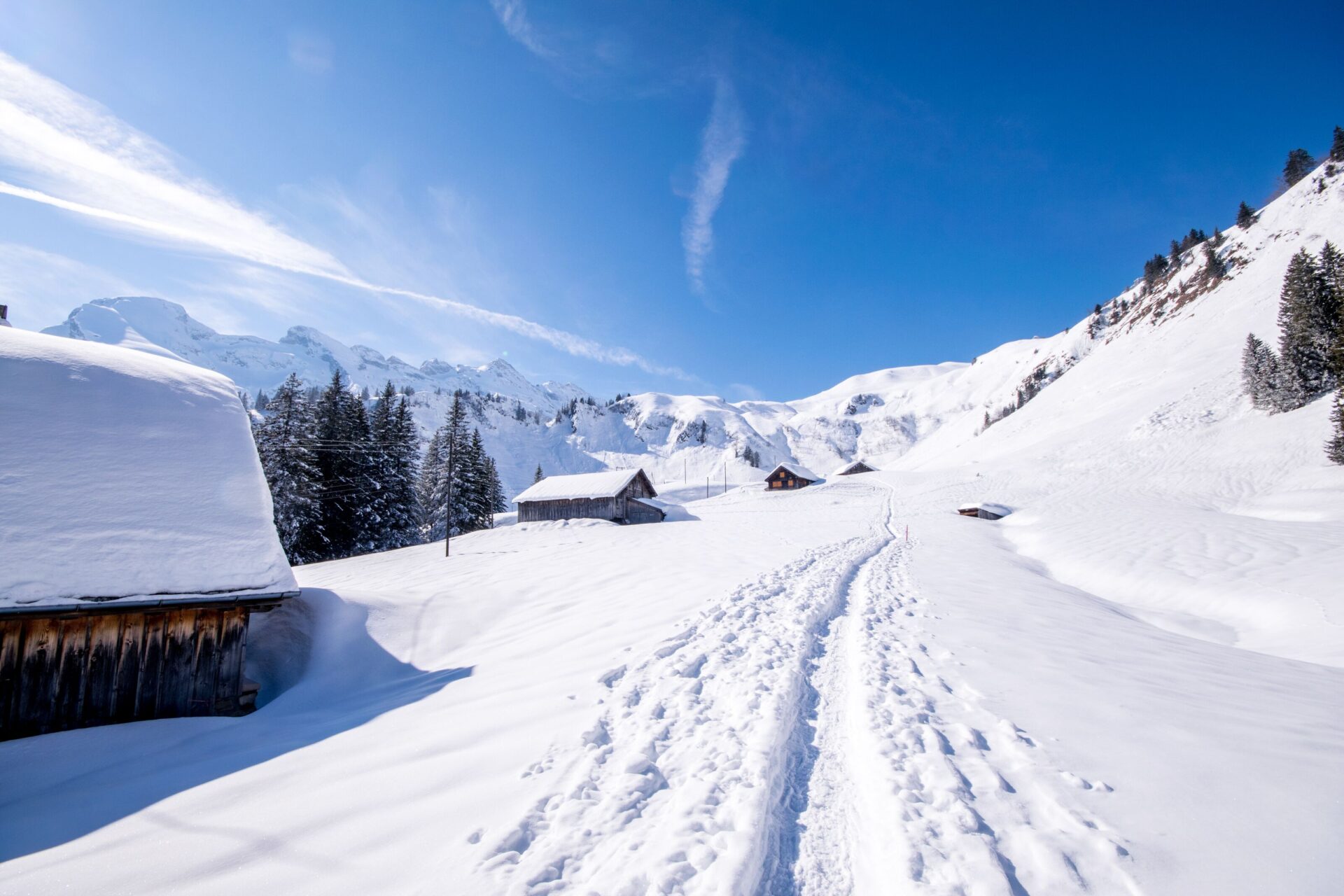 Eine tief verschneite Winterlandschaft mit Spuren auf einem Pfad führt vorbei an verschneiten Hütten, umgeben von hohen Bergen unter strahlend blauem Himmel. Die ruhige Szene vermittelt das Gefühl von Abgeschiedenheit und natürlicher Schönheit.