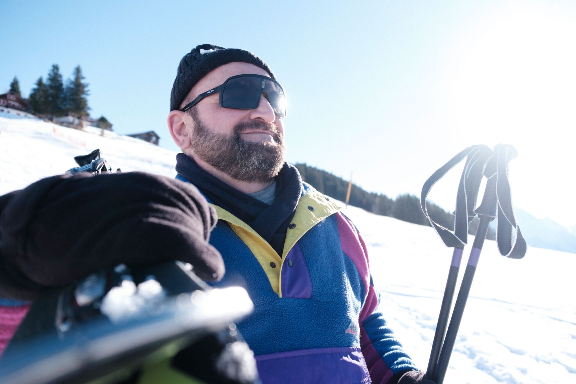 Ein lächelnder Mann mit Sonnenbrille und bunter Skijacke steht auf einer verschneiten Piste, während die Sonne hell vom Himmel scheint. Im Hintergrund sind Bäume und ein Teil eines Gebäudes zu erkennen, die das winterliche Ambiente abrunden.