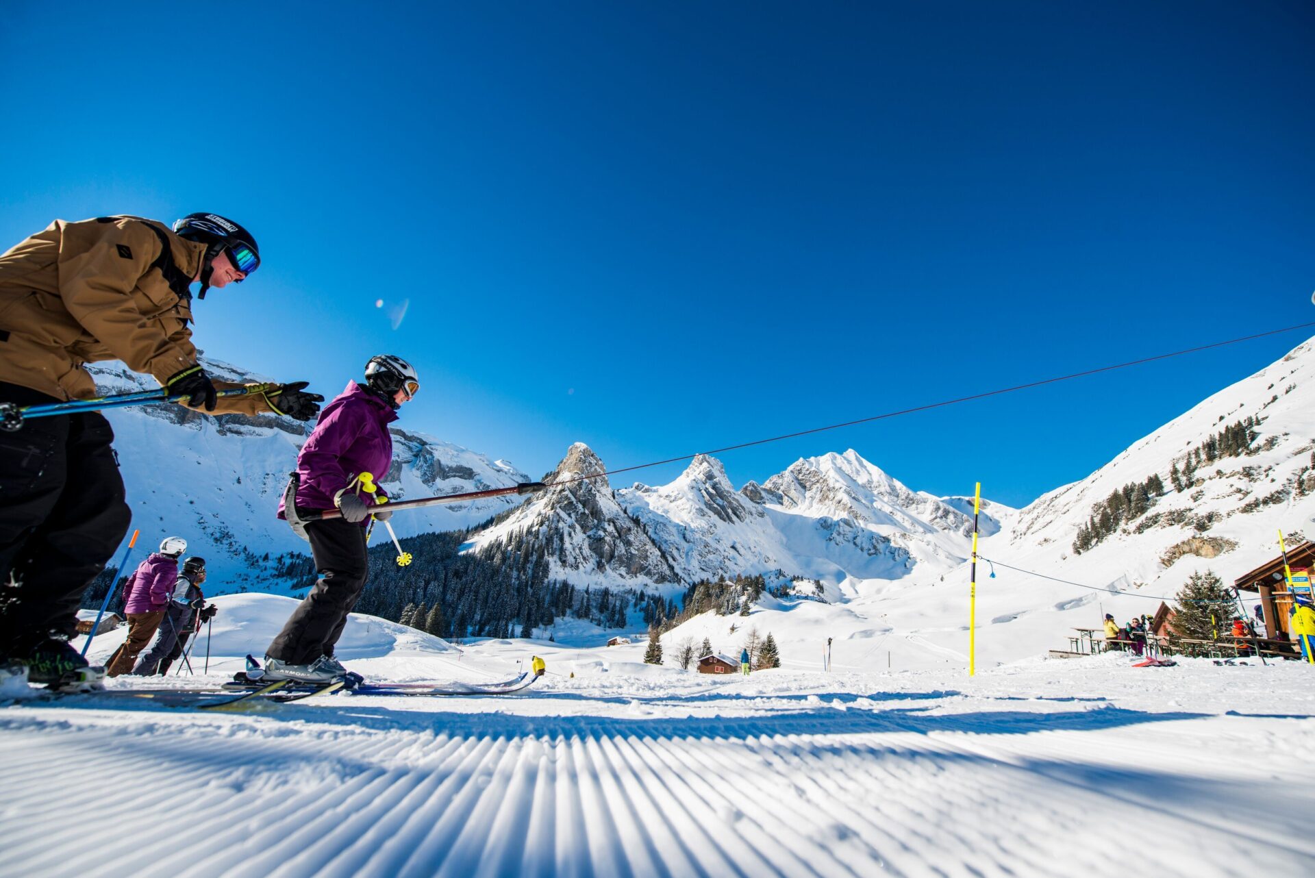 Skifahrer greifen einen Skilift, um den sonnigen Hang hinaufzuziehen, während sich vor ihnen eine beeindruckende Bergkulisse erhebt. Die klare Luft und das strahlende Wetter schaffen eine dynamische und motivierende Wintersportatmosphäre.