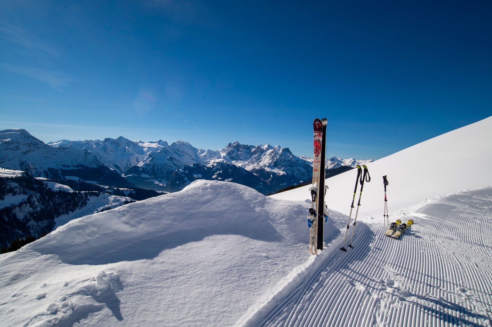 Zwei Paar Skier und Skistöcke stehen im tiefen Schnee an einer präparierten Piste, während sich im Hintergrund eine atemberaubende Berglandschaft erstreckt. Die klare Winteratmosphäre und das weite Panorama laden zum Skifahren ein.