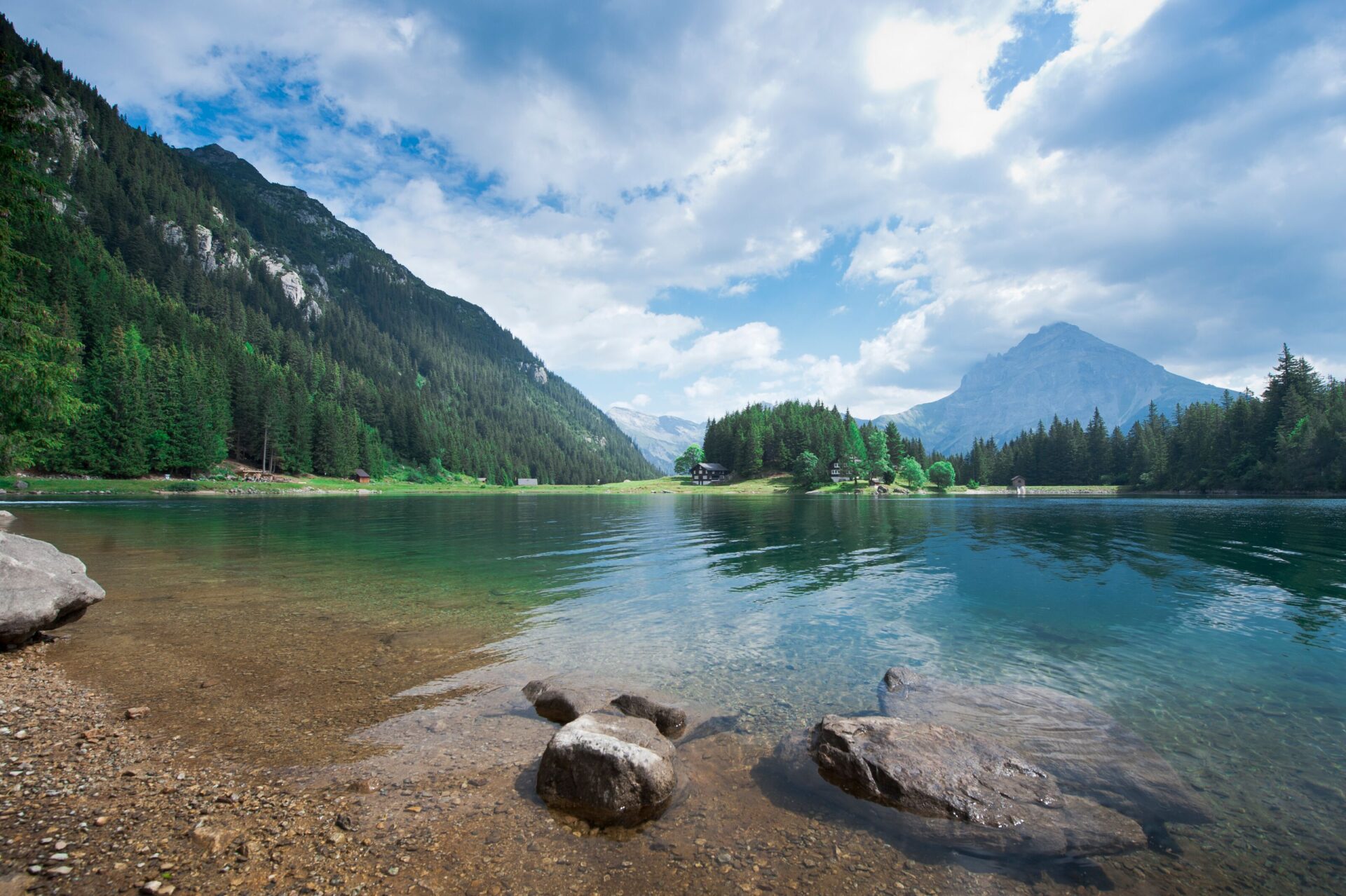 Das Bild zeigt einen klaren Bergsee, dessen Ufer von runden Steinen und grünen Wäldern umgeben ist, während im Hintergrund majestätische Berge in den Himmel ragen. Die Spiegelung des Himmels und der Wolken auf der ruhigen Wasseroberfläche verleiht der Szene eine friedliche Stimmung.