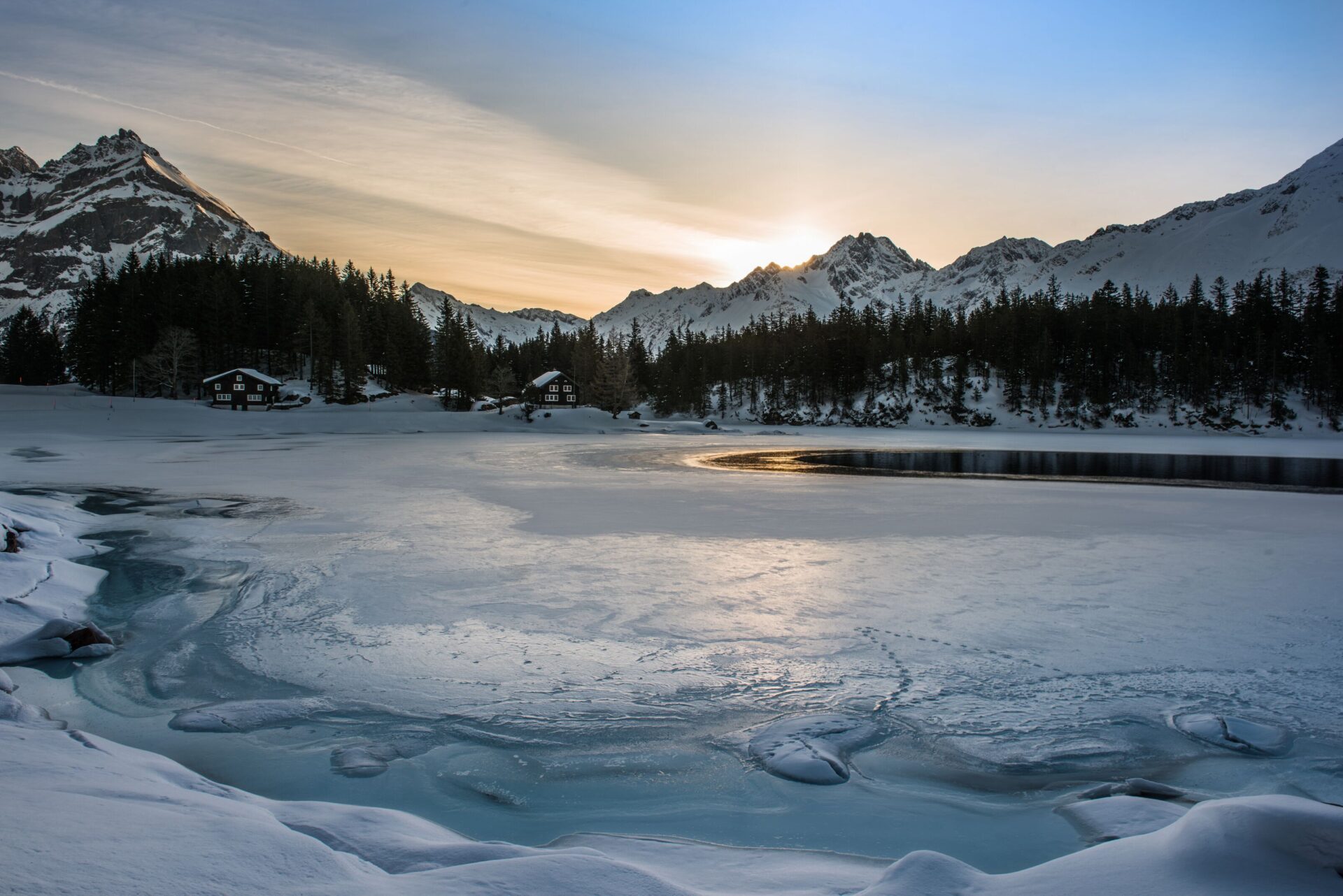 Die untergehende Sonne taucht einen zugefrorenen Bergsee in sanftes Licht, umgeben von schneebedeckten Bäumen und Gipfeln. Die Szene strahlt eine ruhige, winterliche Abendstimmung aus.