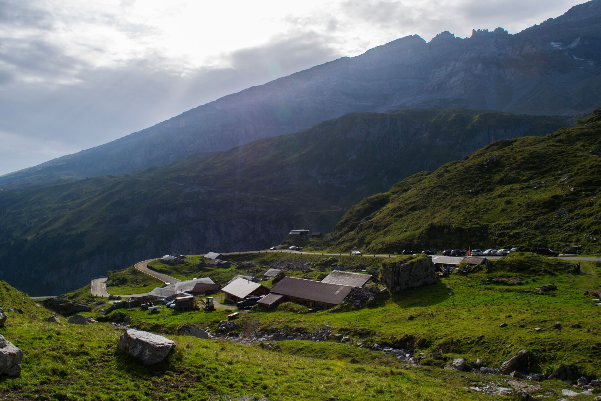 Eine kleine Ansammlung von Berghütten liegt inmitten grüner Hänge, umgeben von hohen Bergketten, während die Sonne durch die Wolken bricht und die Landschaft sanft beleuchtet. Die Szene vermittelt Ruhe und die Abgeschiedenheit des Berglebens.