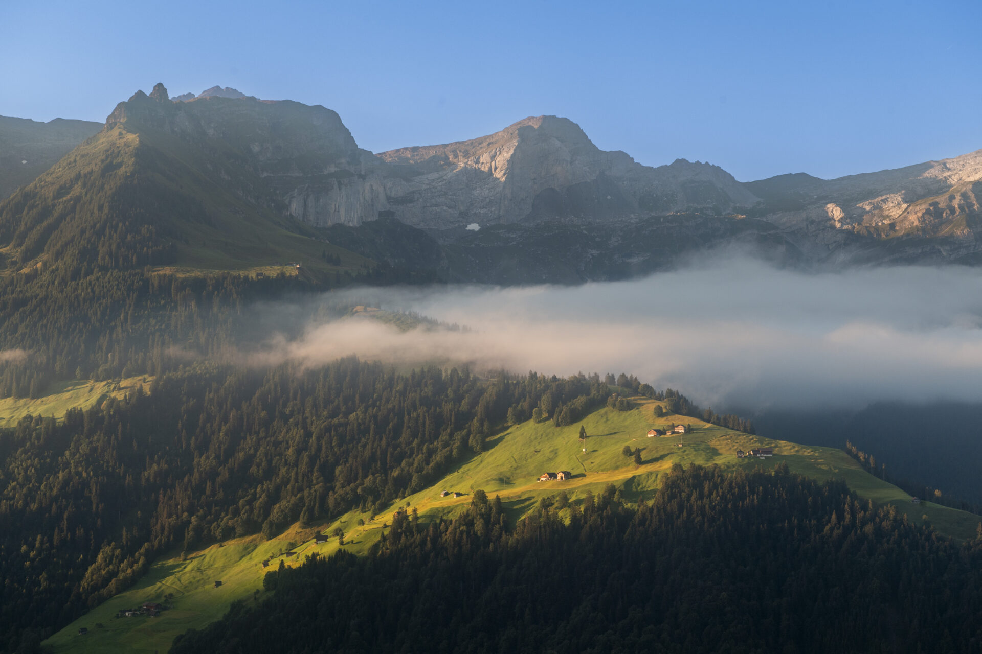 Ein nebelverhangenes Tal mit grünen Wiesen und verstreuten Berghütten liegt unter imposanten Felswänden, die im weichen Morgenlicht erstrahlen. Die Szenerie wirkt ruhig und mystisch und zeigt die stille Schönheit der Berglandschaft.