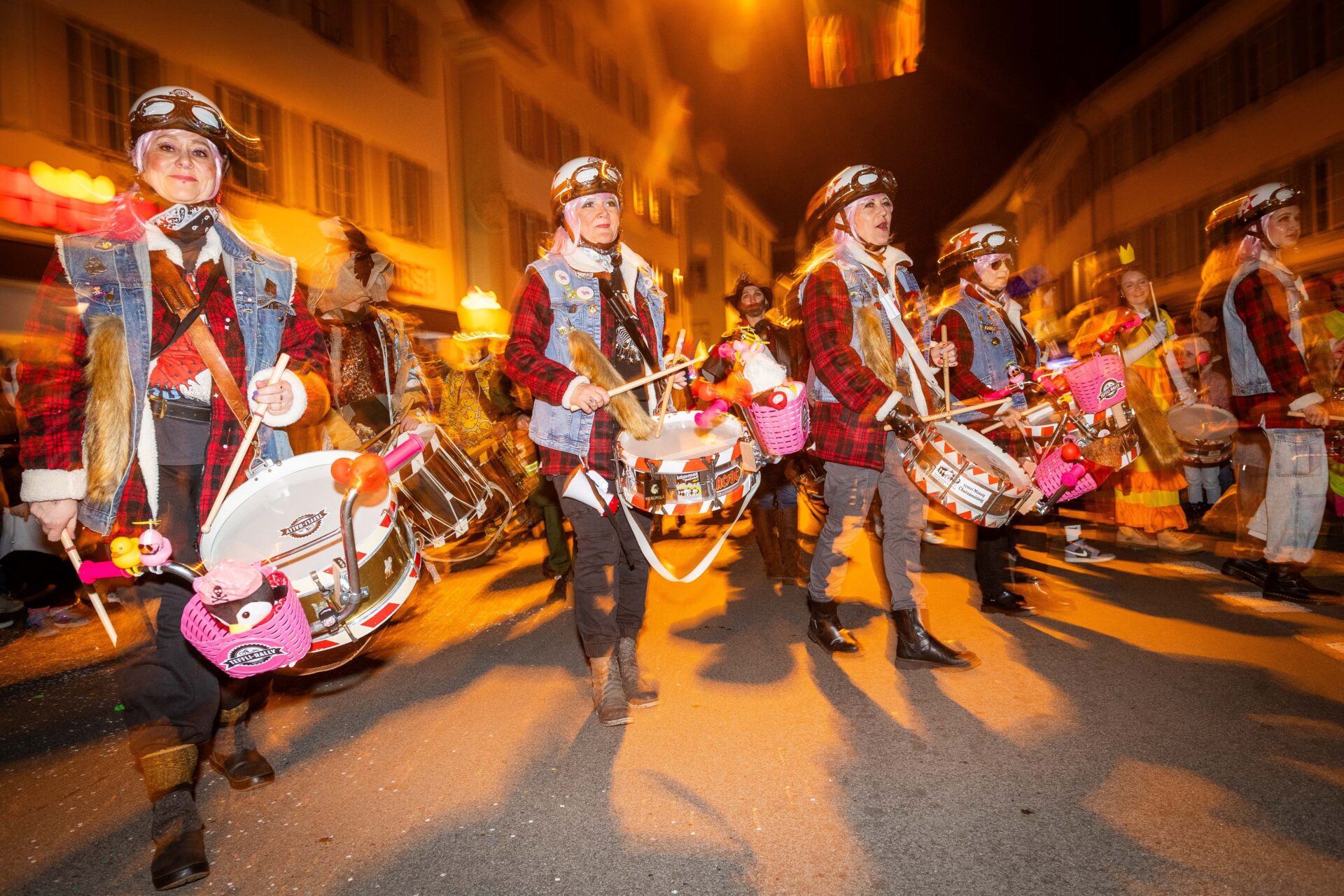 Eine Trommlergruppe in detailreichen, historischen Uniformen marschiert mit leuchtend pinken Trommeln durch die nächtlichen Straßen. Die warmen Lichter der Stadt und die bewegte Szenerie unterstreichen die dynamische und festliche Atmosphäre des Karnevalsumzugs.