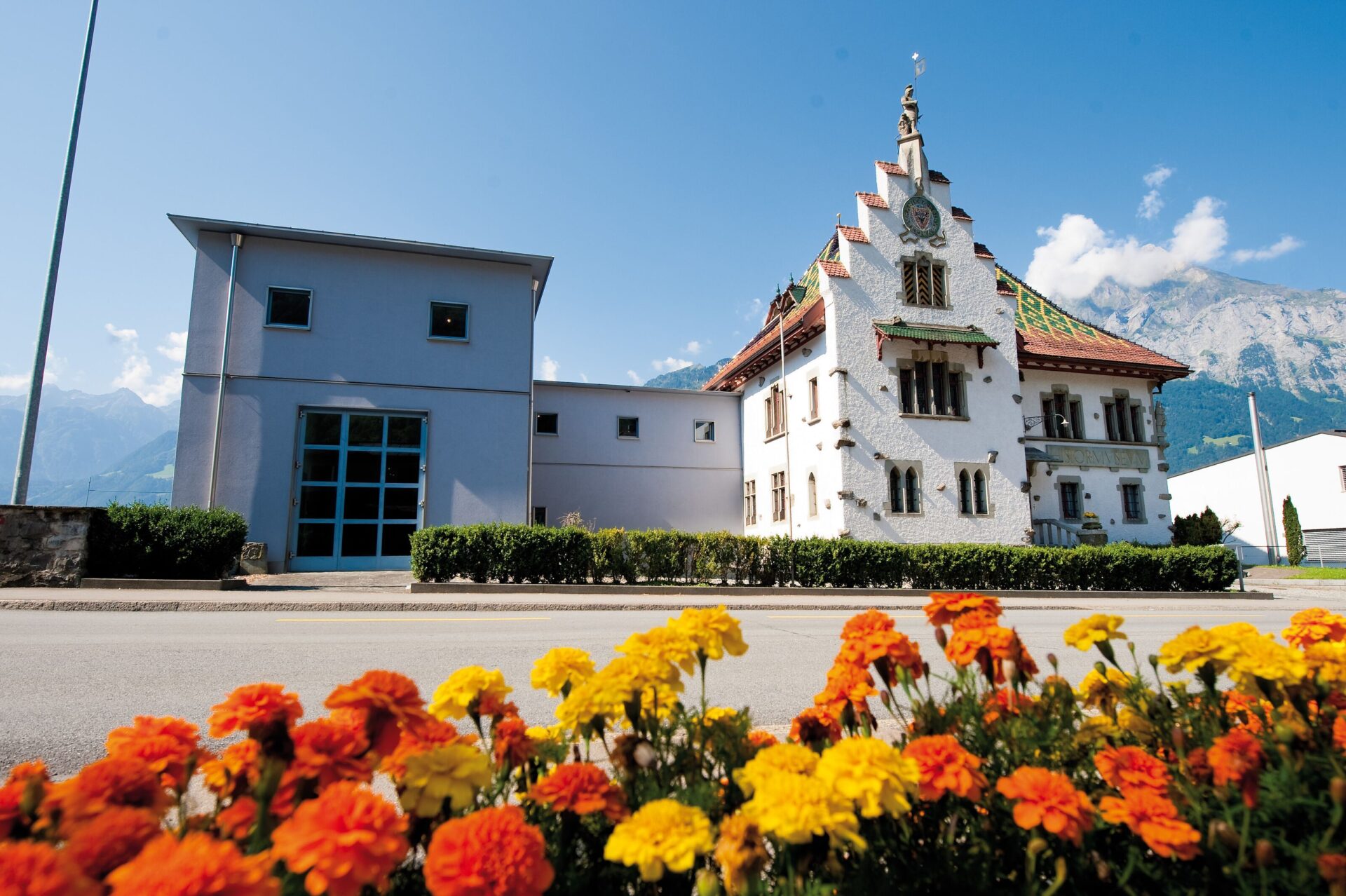 Ein historisches Gebäude mit verzierten Giebeln steht neben einem modernen Anbau, während im Vordergrund bunte Blumen blühen und Berge im Hintergrund aufragen. Die Szene zeigt den Kontrast zwischen Tradition und Moderne in einer malerischen Umgebung.