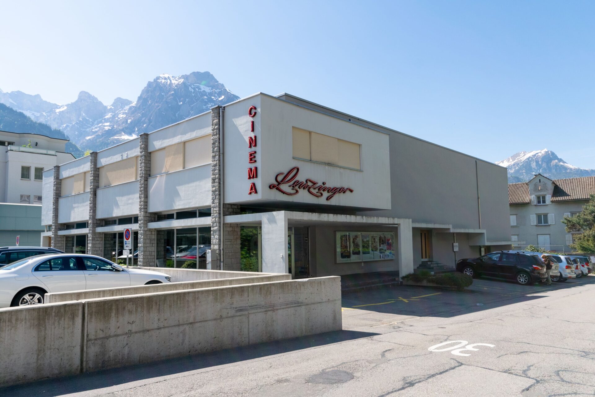 Ein kleines Kino mit dem Namen „Cinema Leuzinger“ steht in einem modernen Gebäude vor einer Kulisse schneebedeckter Berge. Die Szene zeigt eine Kombination aus städtischer Architektur und alpiner Umgebung.