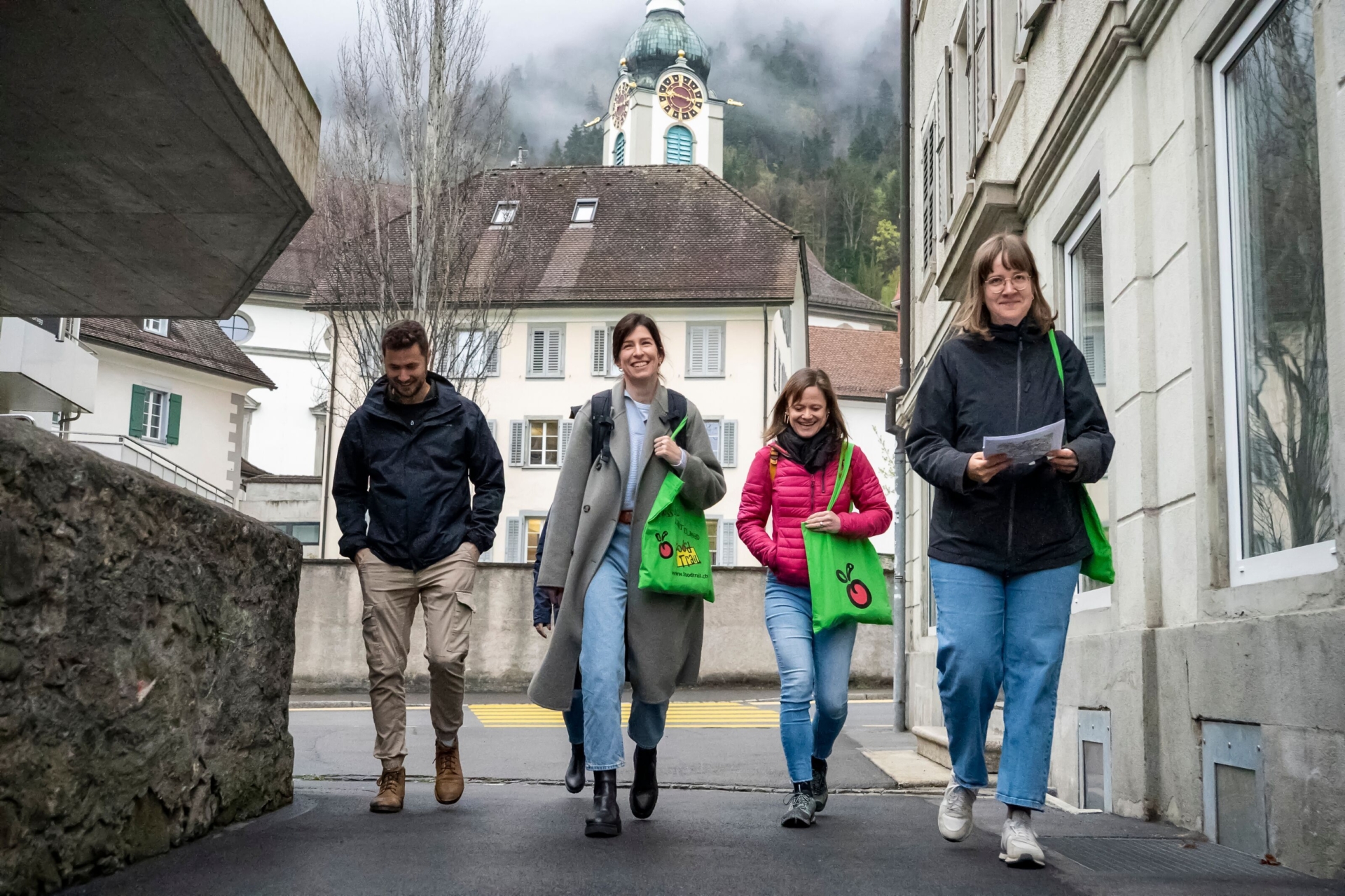Vier Personen gehen lächelnd durch eine enge Straße eines Dorfes, während sie grüne Taschen und eine Karte in der Hand tragen. Im Hintergrund ragt eine Kirche mit Turmuhr vor nebelverhangenen Bäumen auf und verleiht der Szene eine ruhige, leicht mystische Stimmung.