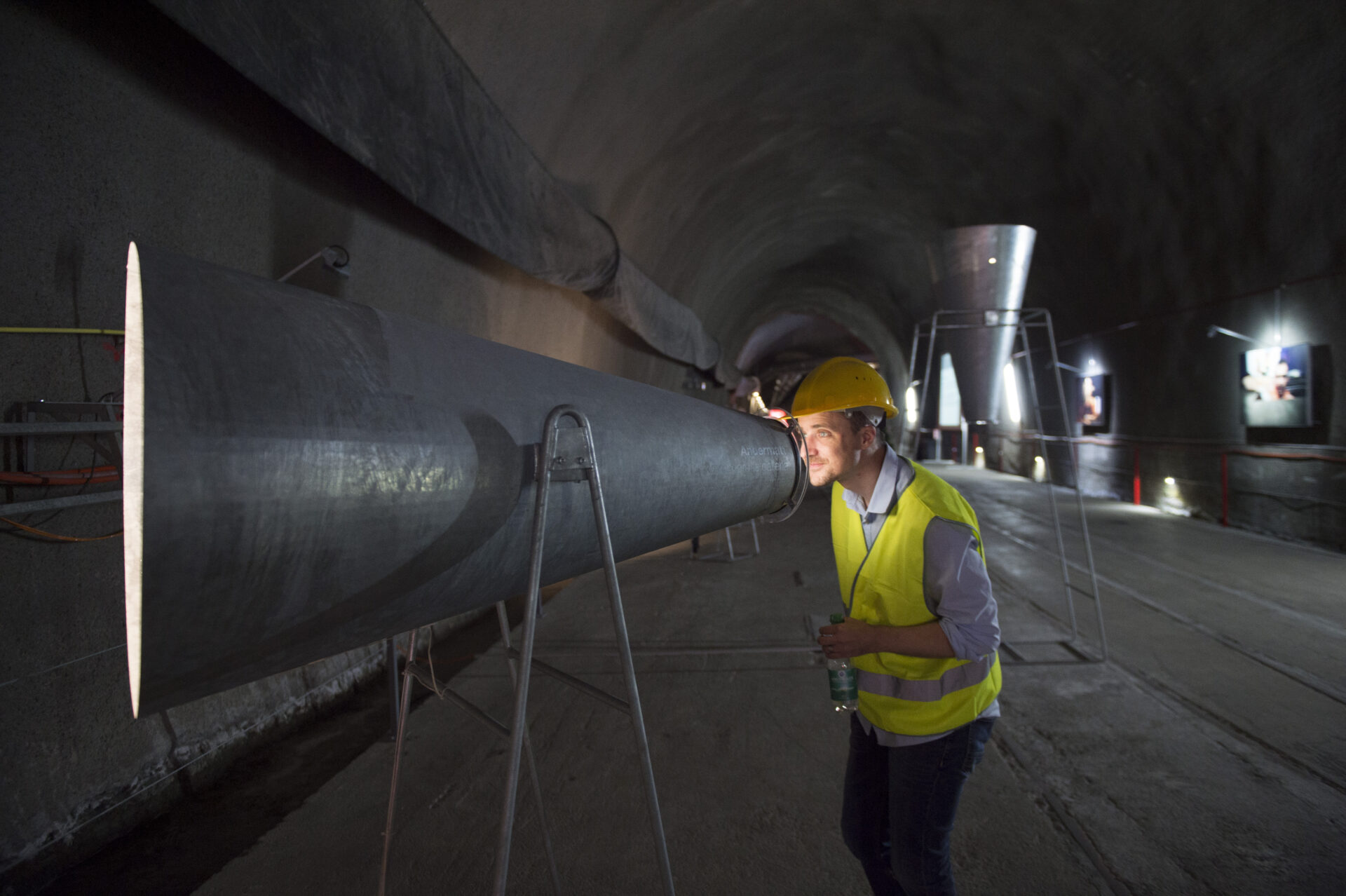 Eine Person in Schutzhelm und Warnweste betrachtet aufmerksam eine große Röhre in einem weitläufigen Tunnel. Die Szene wirkt industriell und erweckt Neugier, mit einer Beleuchtung, die den Tunnel strukturiert und tief wirken lässt.