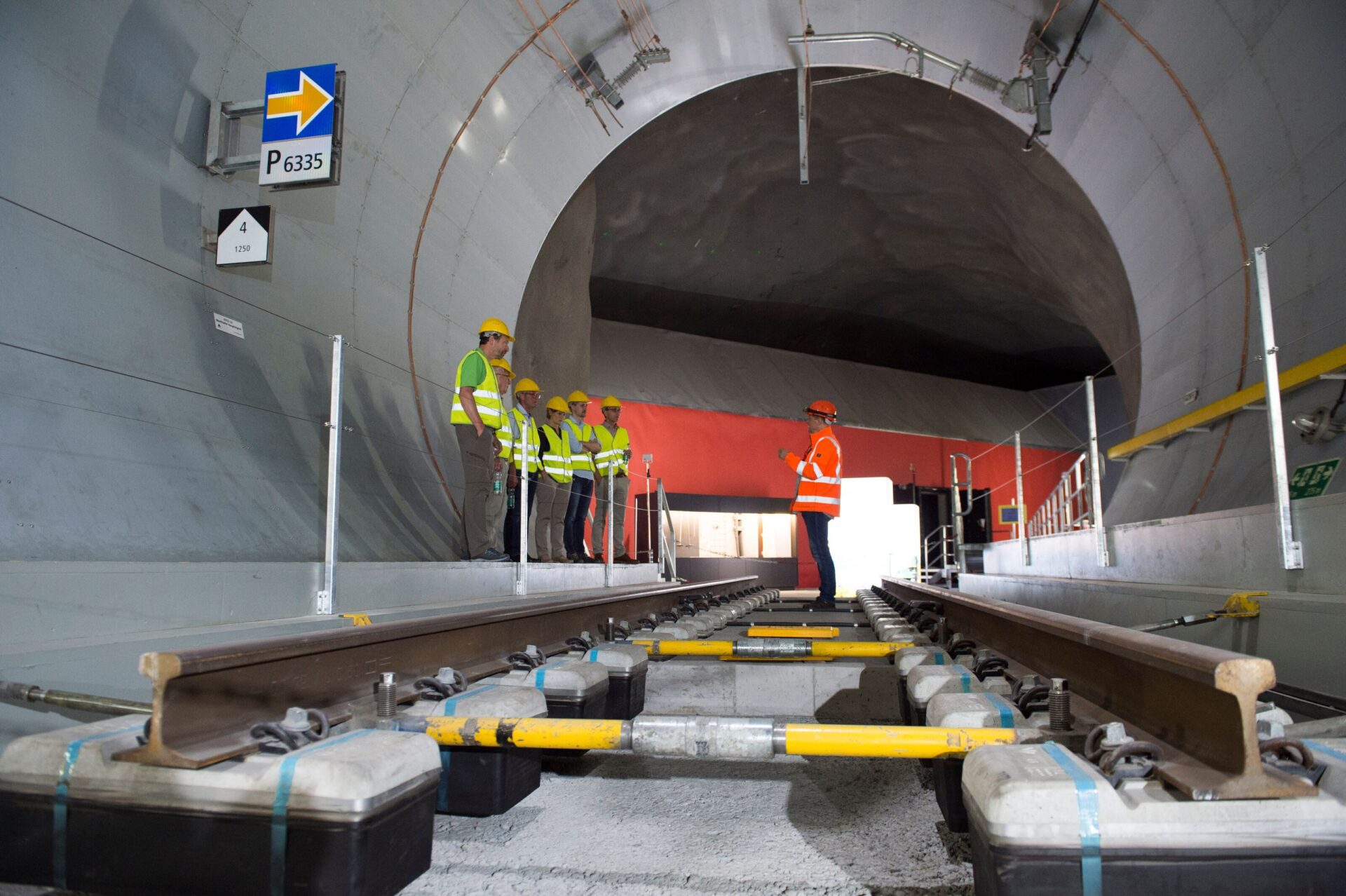 Eine Gruppe von Menschen in Warnwesten und Schutzhelmen steht am Eingang eines modernen Eisenbahntunnels, während eine Person in orangefarbener Weste den Tunnel erklärt. Die Szene vermittelt ein Gefühl von Fortschritt und Teamarbeit in einem technischen Umfeld.