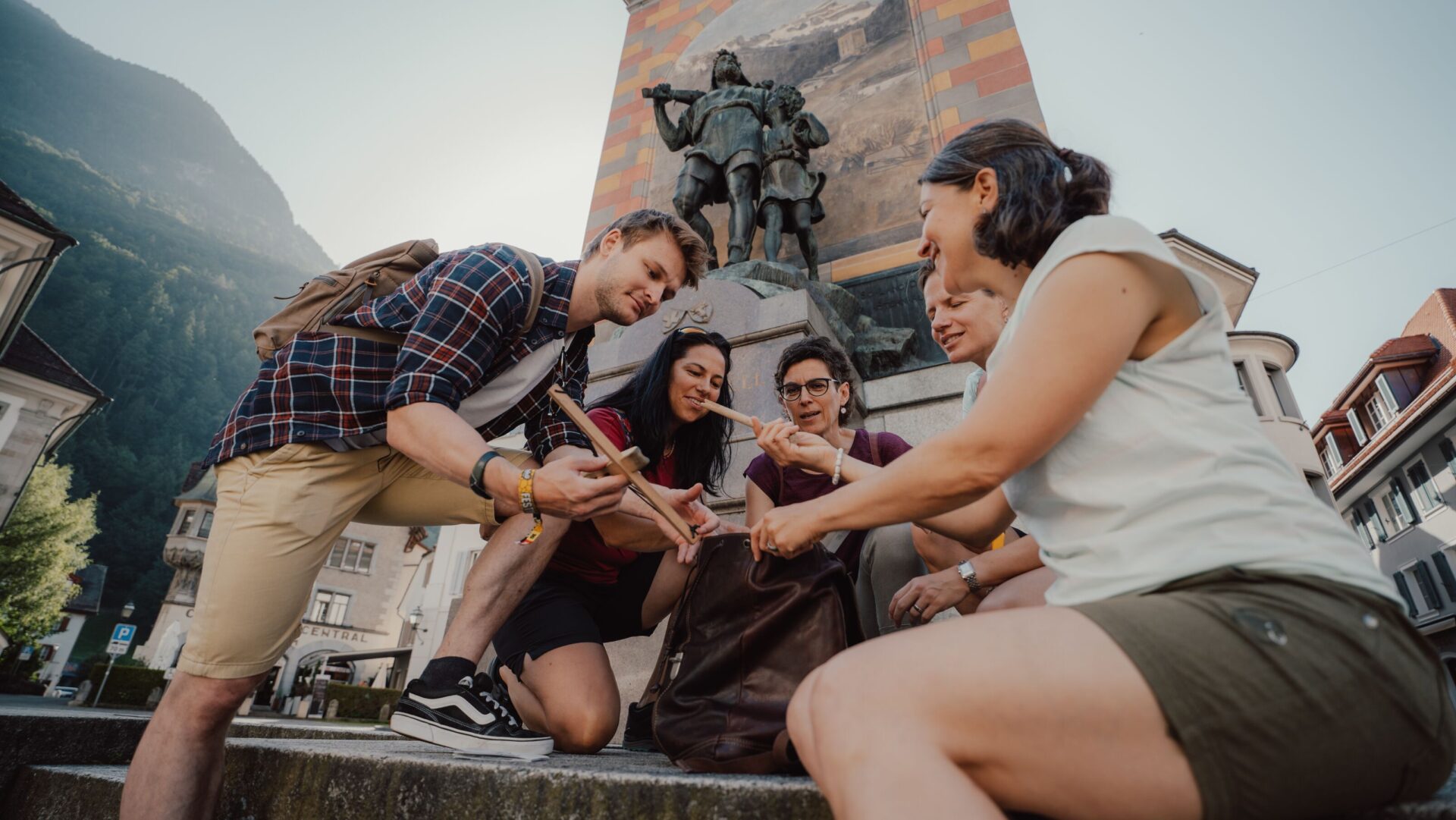 Eine Gruppe von Freunden versammelt sich lächelnd um eine Karte oder ein Smartphone vor einem historischen Denkmal in einer Bergstadt. Die Szene vermittelt Abenteuerlust und die Freude am gemeinsamen Erkunden.