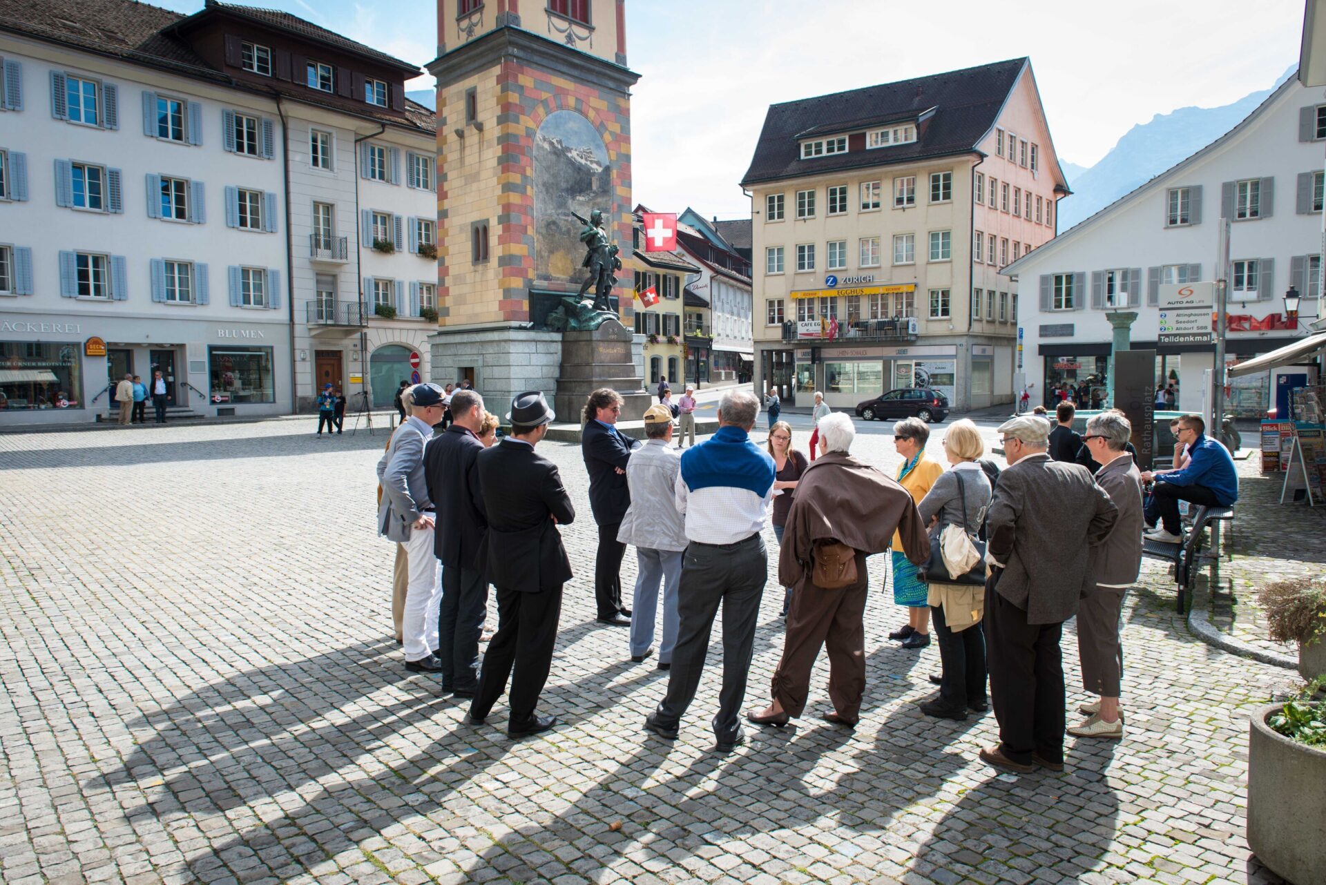 Eine Gruppe von Menschen steht auf einem historischen Platz und hört einem Stadtführer vor einem bemalten Turm aufmerksam zu. Die Szene vermittelt ein Gefühl von Gemeinschaft und kulturellem Interesse in einem malerischen Altstadtambiente.