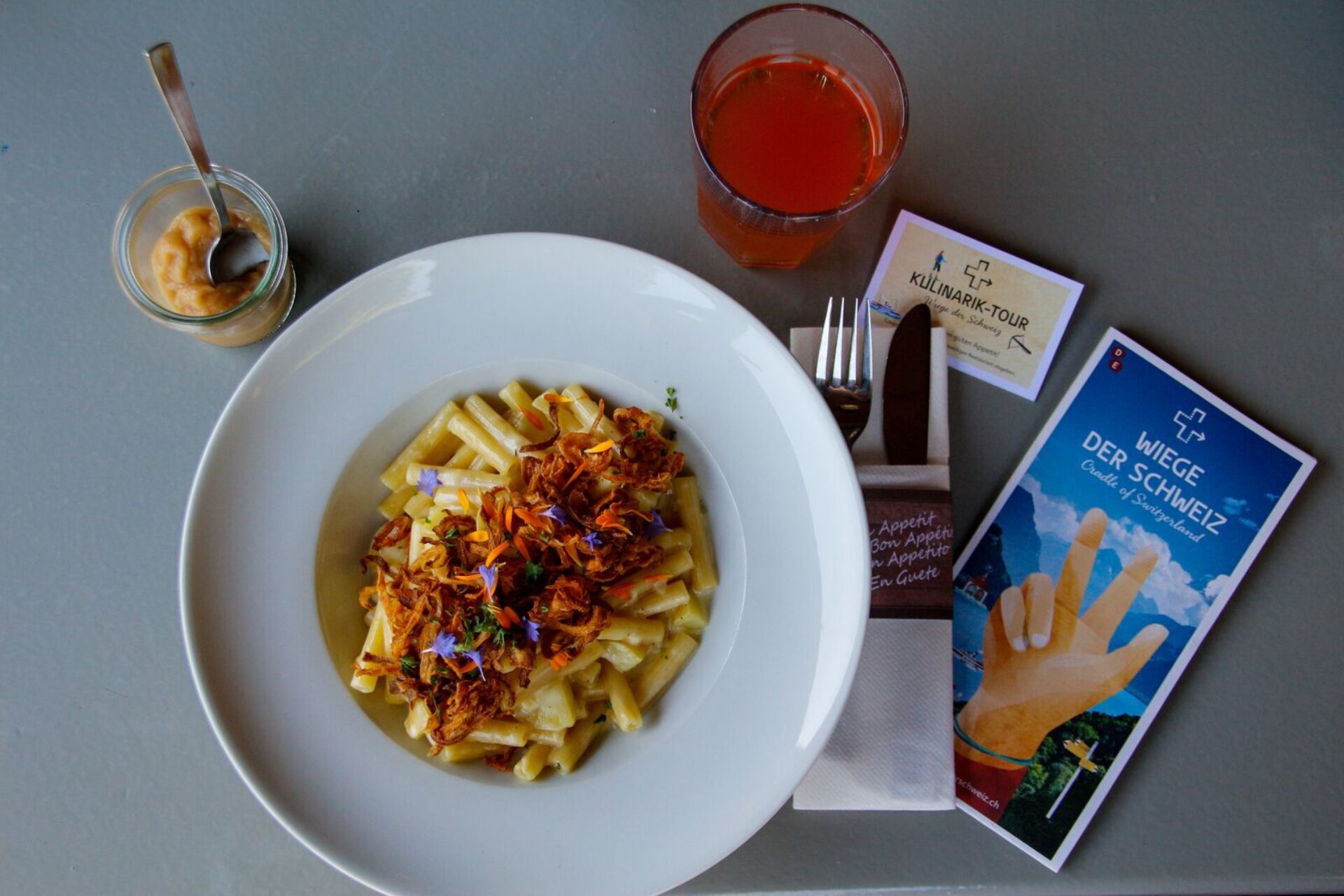 Ein Teller mit Älplermagronen, garniert mit Röstzwiebeln und Blüten, steht neben einem Glas rotem Saft, einem kleinen Dessert im Glas sowie Informationsmaterial über die Region. Die liebevoll angerichtete Mahlzeit verbindet alpine Tradition mit Genuss in modernem Ambiente.