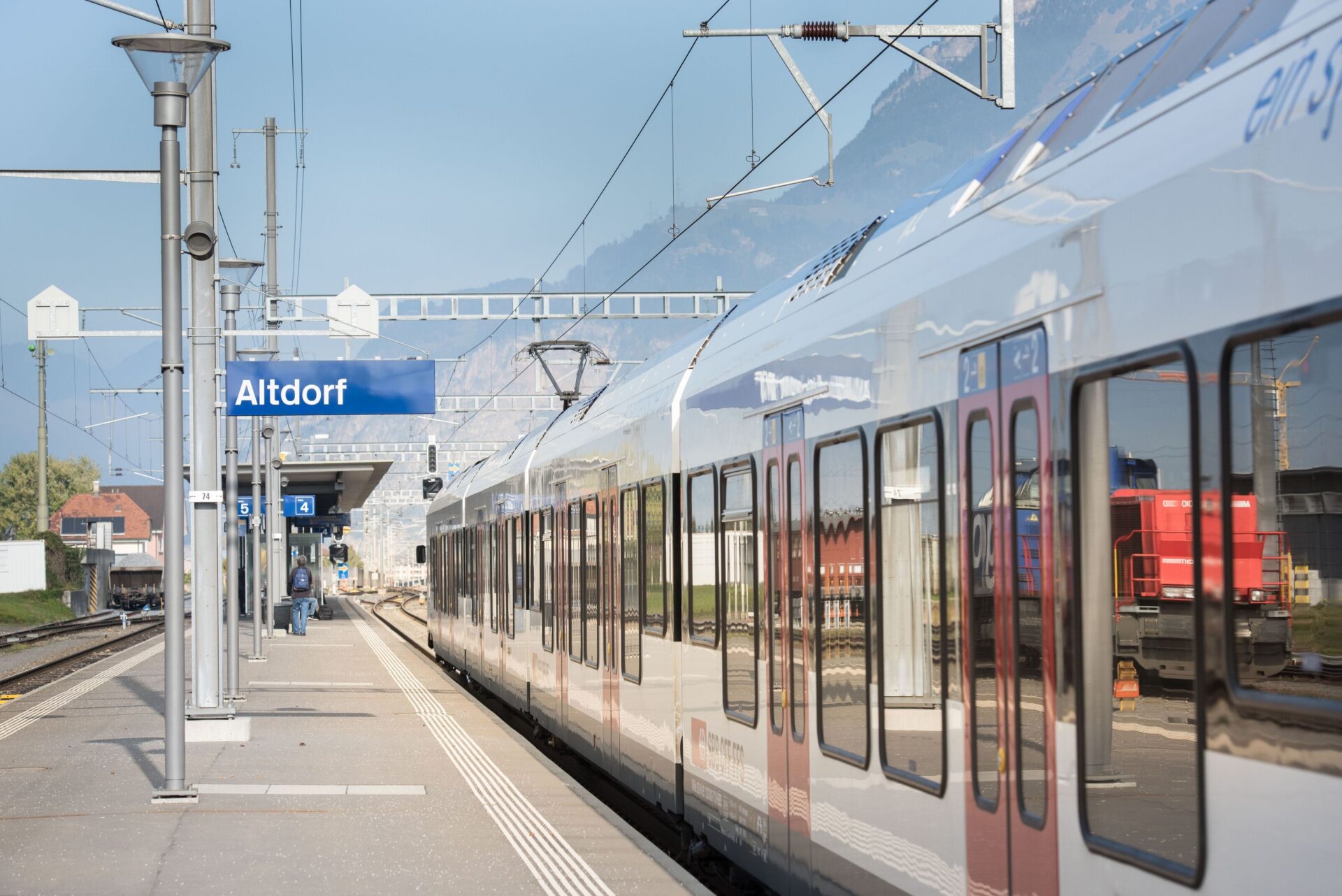 Ein moderner Zug steht am Bahnhof Altdorf, wo sich die Bahnsteigschilder und die klare Architektur gegen den blauen Himmel abheben. Die Szene vermittelt einen Eindruck von moderner Infrastruktur und Mobilität in einer malerischen Umgebung.