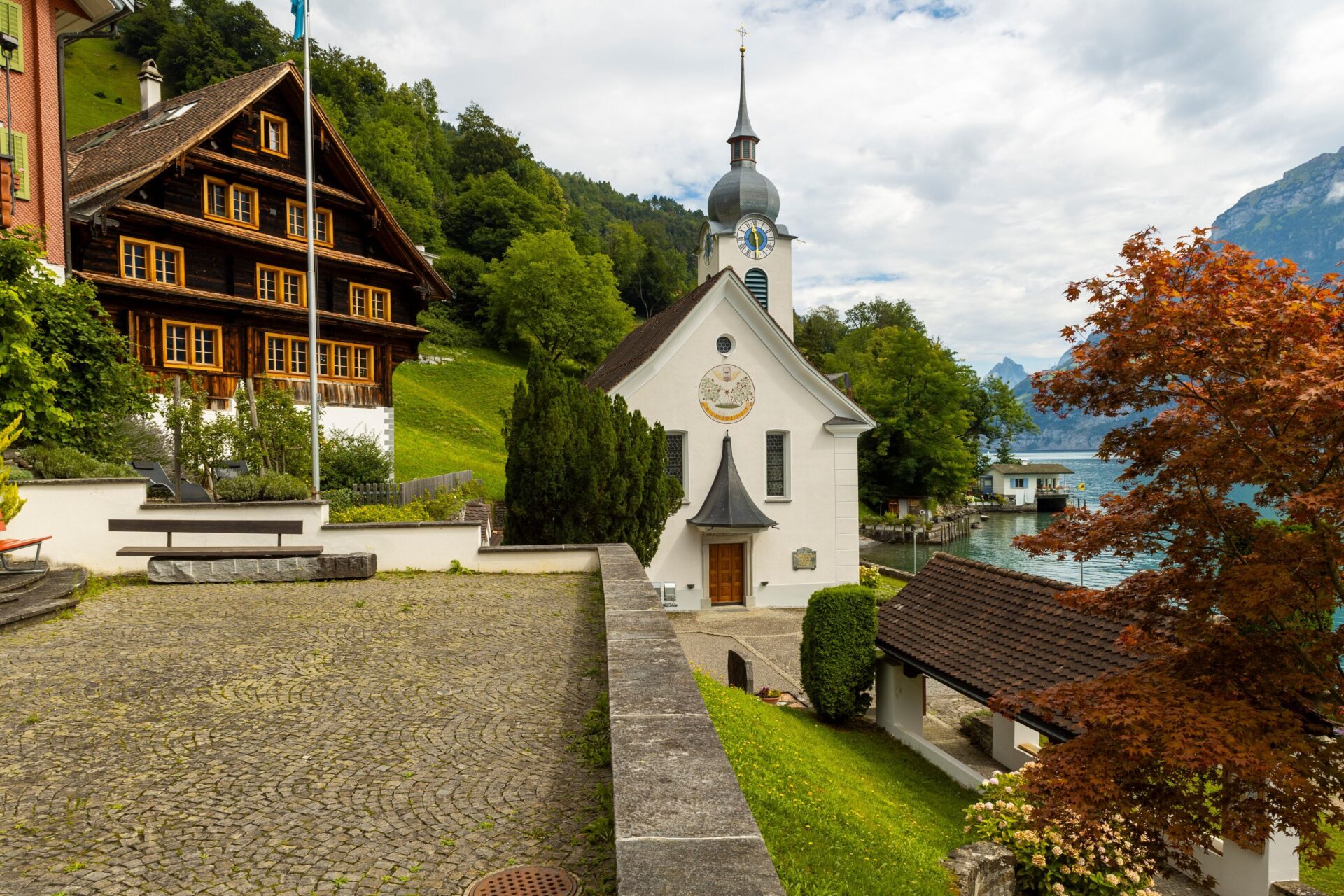 Die malerische Kapelle in Bauen steht vor traditionellen Holzhäusern und bietet einen Blick auf den nahegelegenen See und die umliegenden Berge. Die Szene strahlt Ruhe und den Charme eines idyllischen Alpendorfes aus.