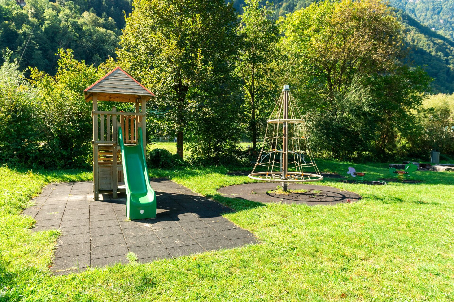 Ein kleiner Spielplatz mit Rutsche und Kletterpyramide liegt eingebettet in eine grüne Wiesenlandschaft vor einer Baum- und Bergkulisse. Die sonnige Umgebung und die Natur schaffen eine einladende und kinderfreundliche Atmosphäre.