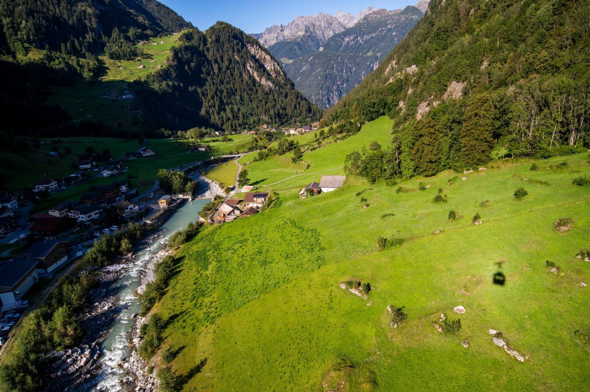 Das Bild zeigt eine idyllische Alpenlandschaft mit saftig grünen Wiesen, einem kleinen Dorf und einem klaren, gewundenen Fluss, der durch das Tal fließt. Im Hintergrund erheben sich majestätische Berge mit felsigen Gipfeln unter einem strahlend blauen Himmel.