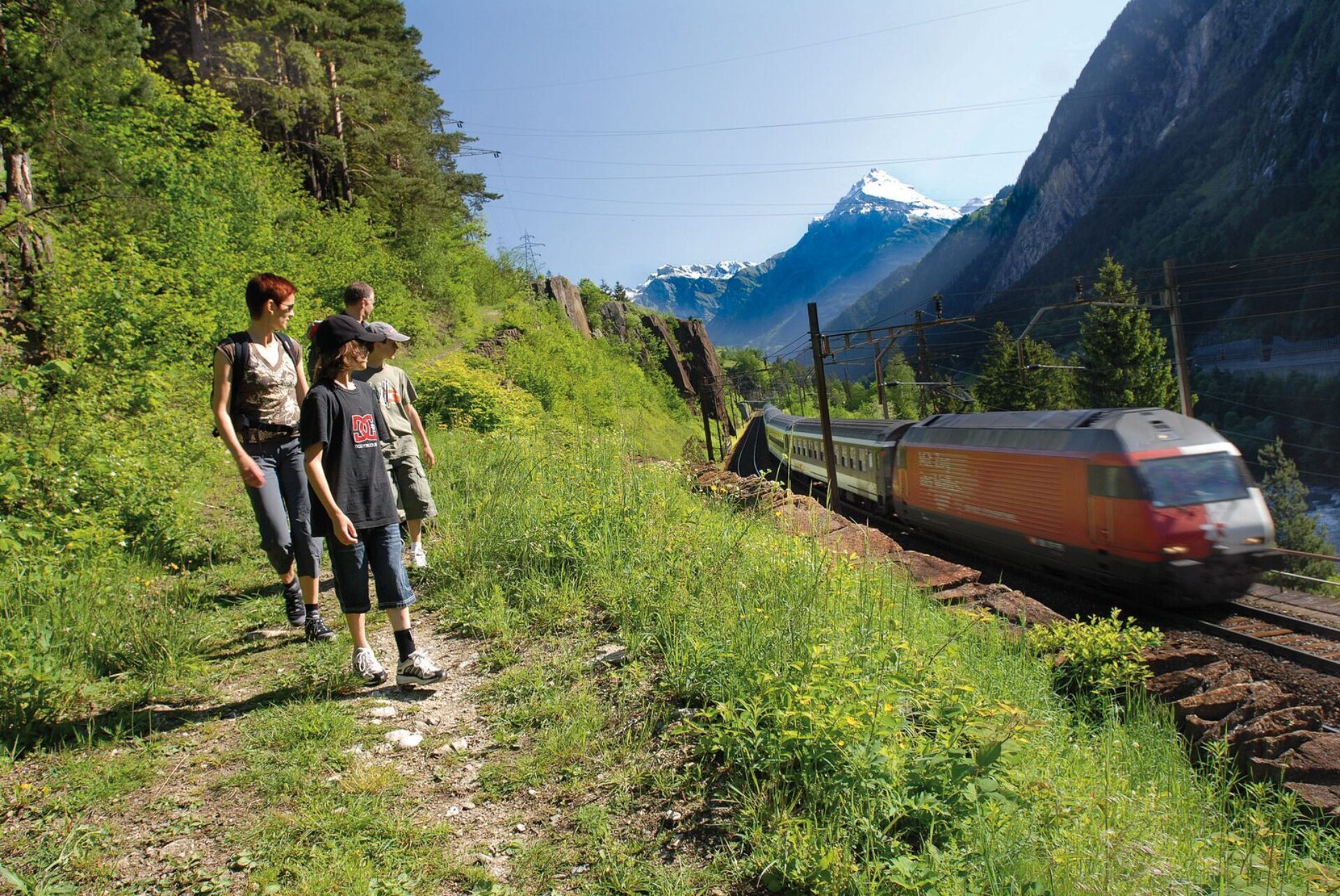 Eine Familie wandert auf einem grünen Pfad entlang einer Bahnstrecke in Silenen, während ein moderner Zug an ihnen vorbeifährt. Die malerische Bergkulisse und das Zusammenspiel von Natur und Technik verleihen der Szene eine dynamische und zugleich friedliche Atmosphäre.