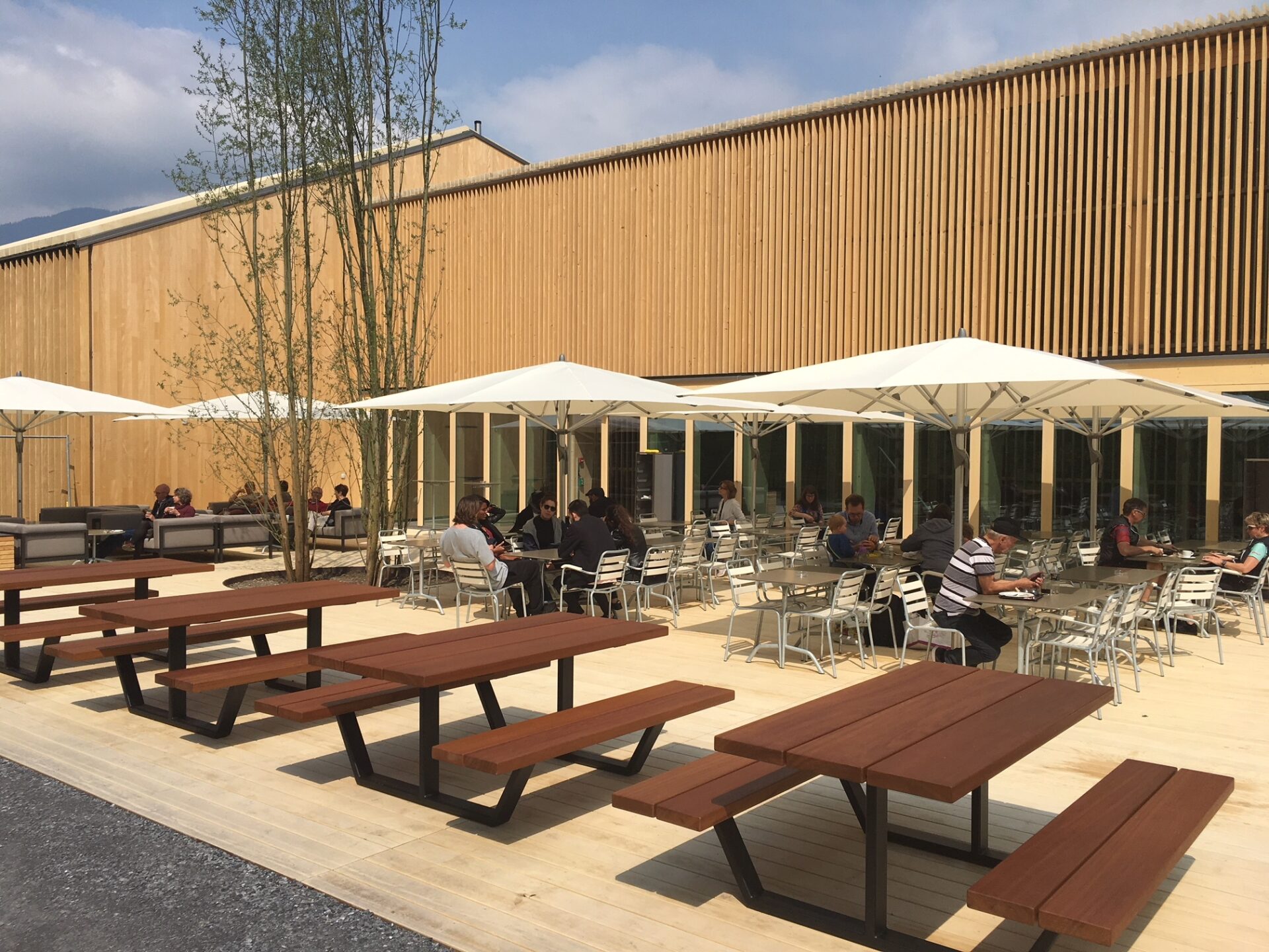 Eine moderne Außenterrasse mit Holzfassade, ausgestattet mit Bänken und Tischen sowie weißen Sonnenschirmen, lädt Besucher zum Verweilen ein. Die helle und offene Gestaltung schafft eine einladende Atmosphäre für entspannte Pausen im Freien.