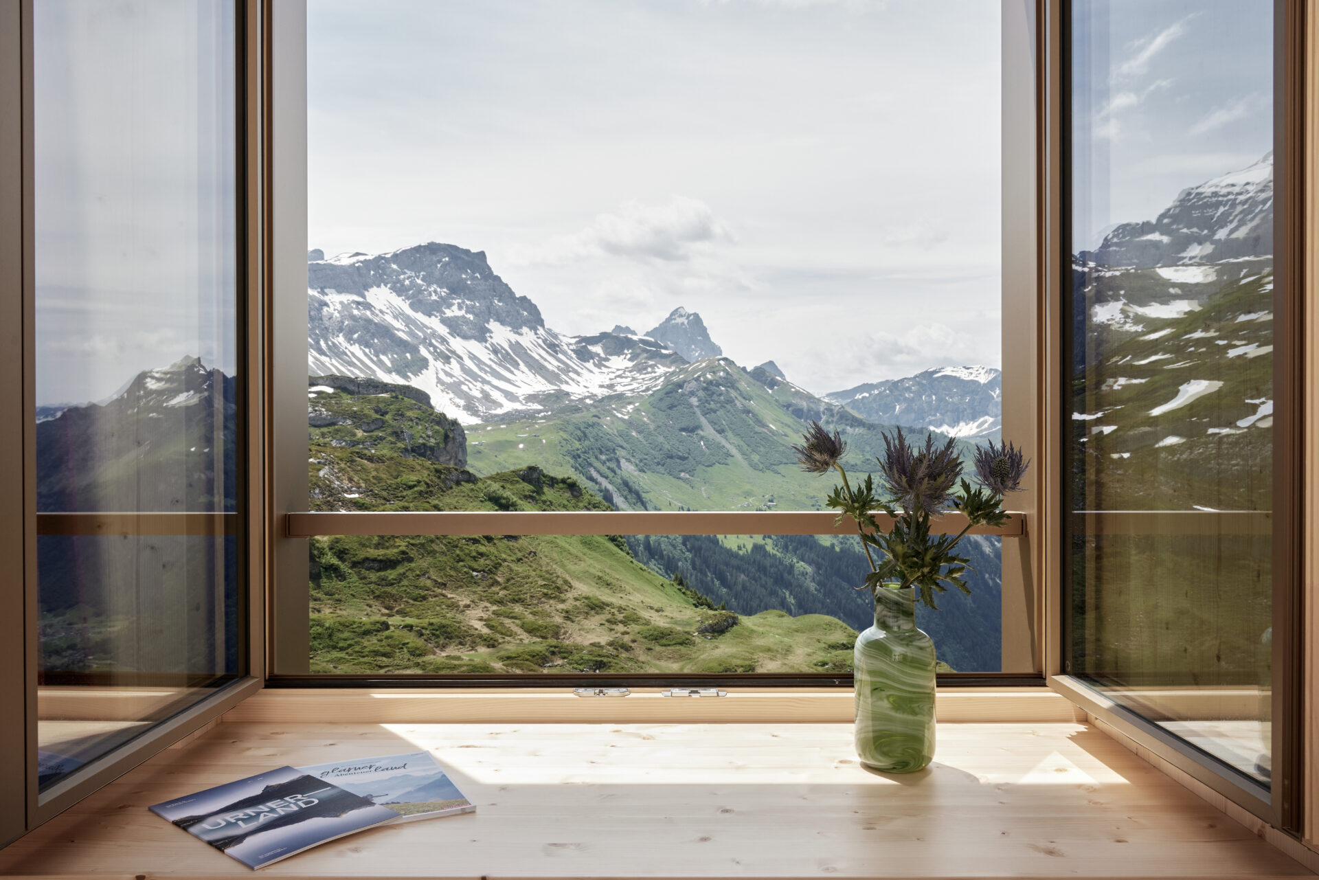 Durch ein offenes Fenster bietet sich ein atemberaubender Blick auf eine grüne Berglandschaft mit schneebedeckten Gipfeln, auf der Fensterbank steht eine Vase mit Blumen. Die Szene vermittelt Ruhe und die Nähe zur Natur, perfekt für eine Auszeit in den Bergen.