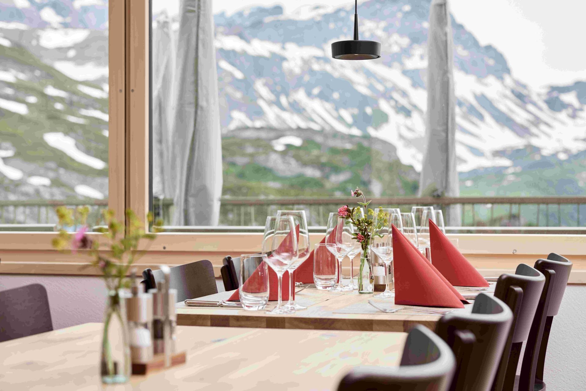 Ein festlich gedeckter Tisch mit roten Stoffservietten und elegantem Geschirr steht vor einem großen Fenster, das den Blick auf eine verschneite Berglandschaft freigibt. Die Kombination aus stilvoller Tischdekoration und alpinem Panorama schafft eine einladende und besondere Atmosphäre.