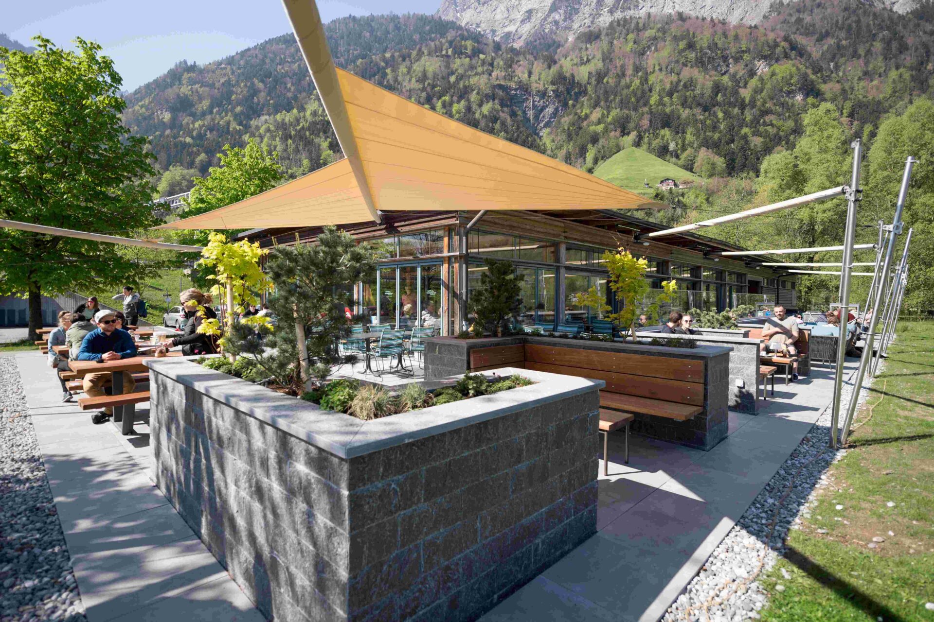 Eine gemütliche Gartenterrasse mit modernen Sitzbereichen, umgeben von Pflanzen und unter schattenspendenden Segeln. Die Kombination aus Natur und moderner Architektur lädt zum Verweilen in entspannter Atmosphäre ein.