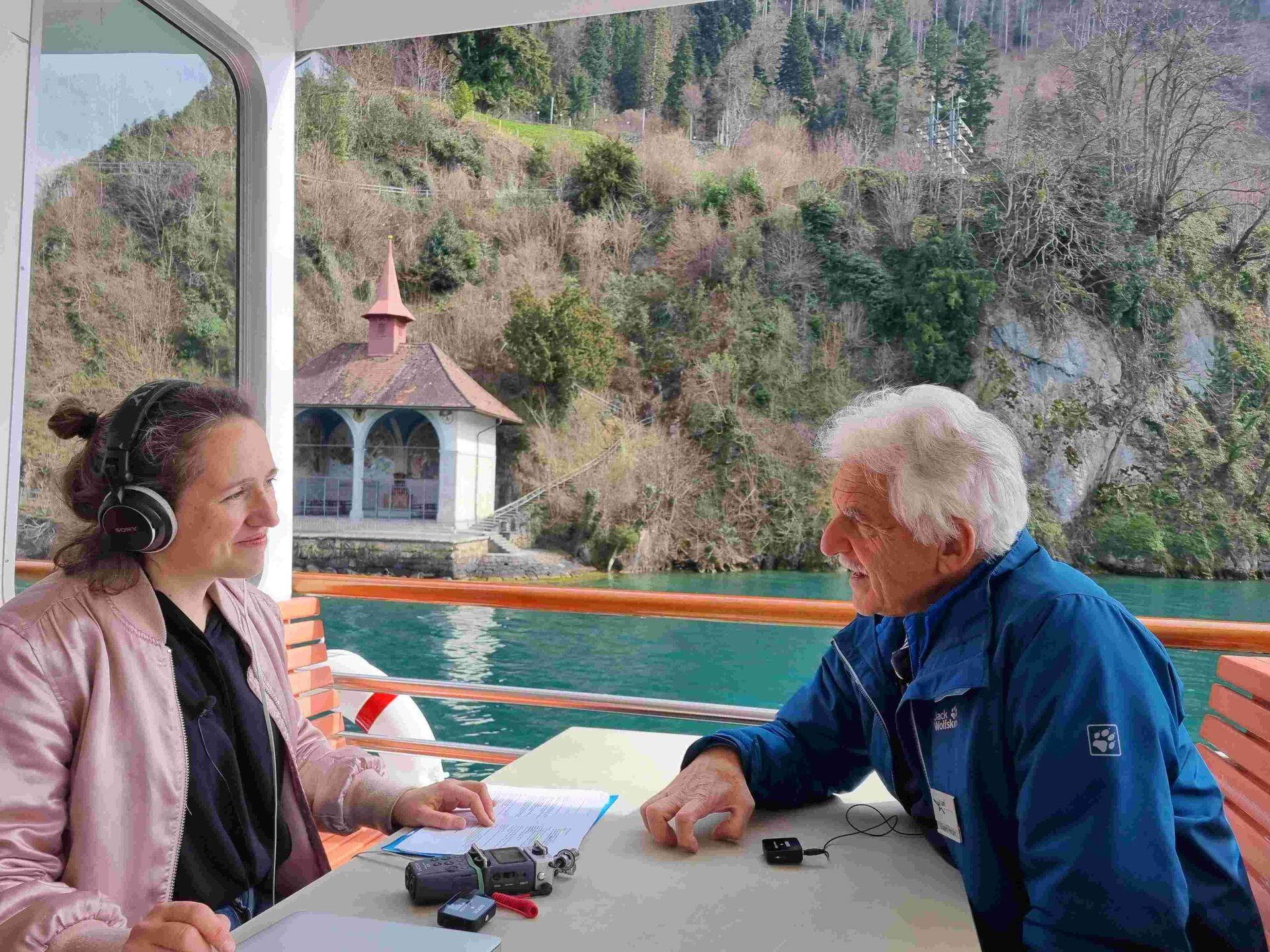 Zwei Personen sitzen sich auf einem Boot gegenüber, eine Person mit Kopfhörern und Aufnahmegerät, die andere im Gespräch vertieft, während im Hintergrund eine idyllische Uferlandschaft vorbeizieht. Die Szene vermittelt eine entspannte Interview-Atmosphäre auf dem Wasser.