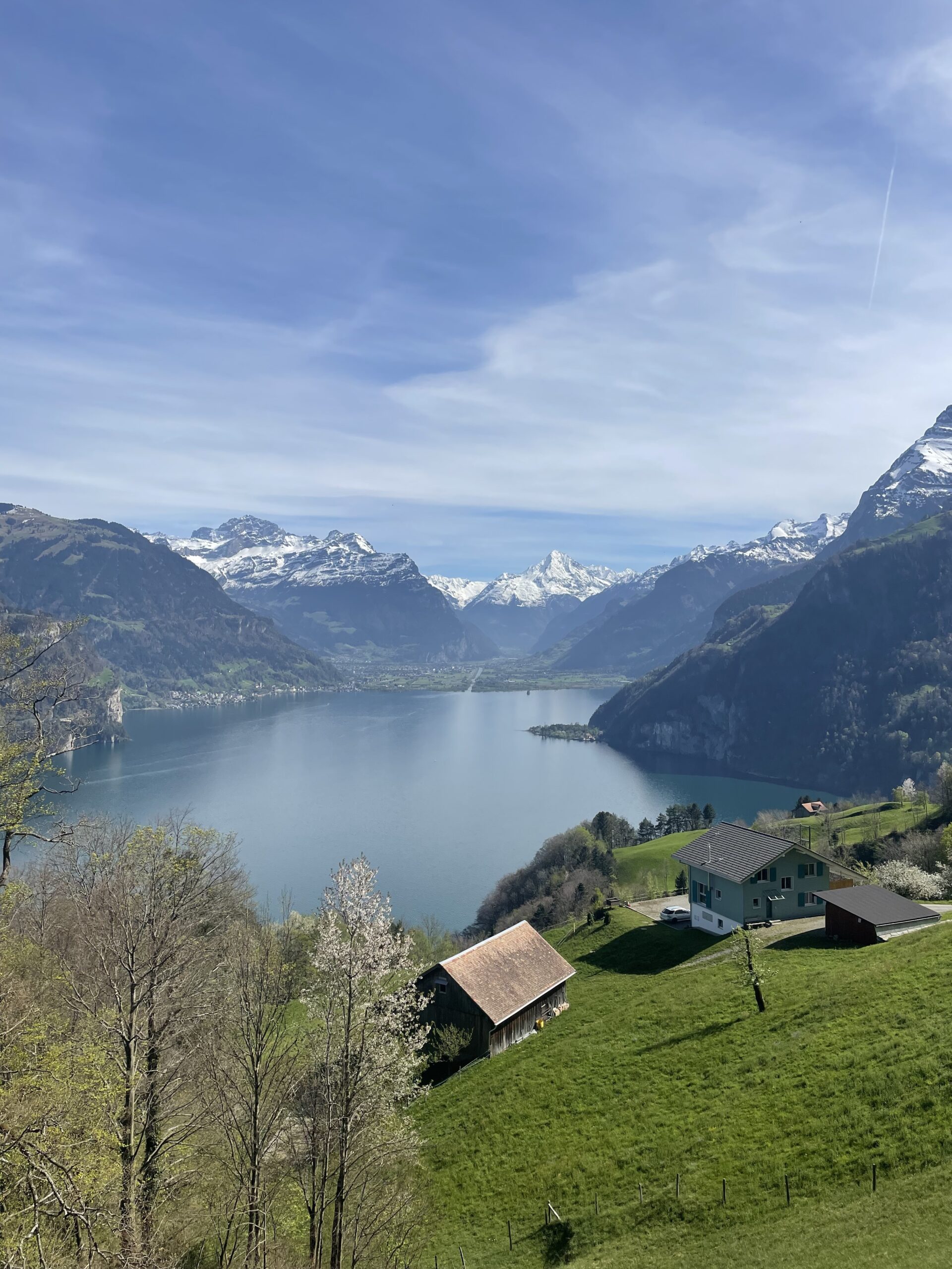 Ein idyllischer Blick auf einen ruhigen Bergsee, umgeben von schneebedeckten Alpen, mit grünen Wiesen und traditionellen Holzhäusern im Vordergrund. Die harmonische Landschaft wirkt friedlich und lädt zum Verweilen ein.