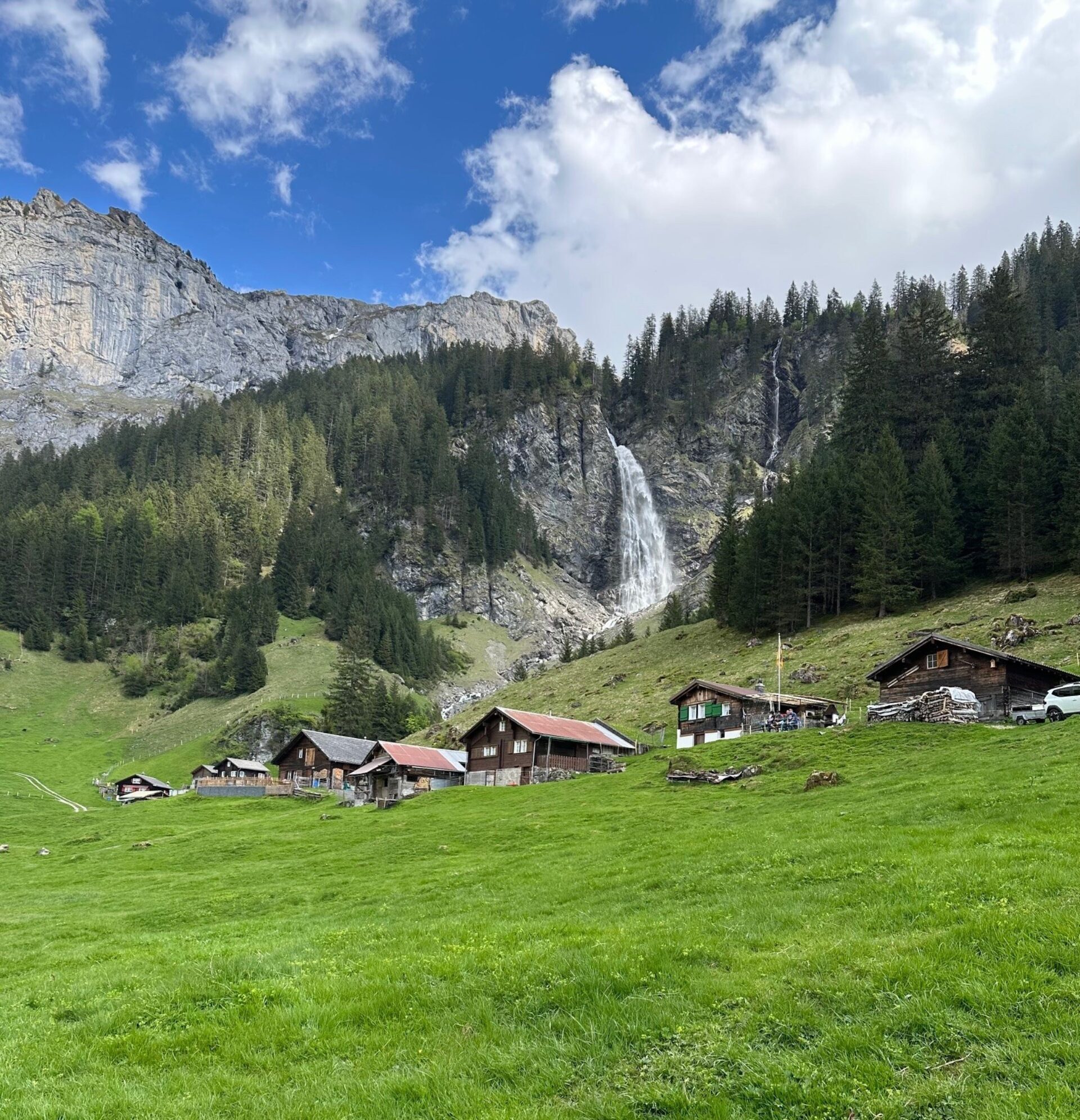 Der Stäubenfall stürzt imposant über eine hohe Felswand ins Tal und bildet eine feine Gischt, die in der Sonne glitzert – ein eindrucksvolles Naturschauspiel inmitten der alpinen Landschaft bei Äsch. Umgeben von sattgrünen Wiesen, Wald und traditionellen Hütten vermittelt die Szenerie Ruhe und Naturgewalt zugleich.