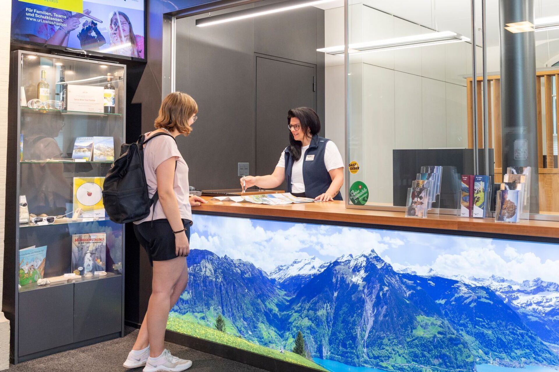 Eine Mitarbeiterin bedient eine Kundin an einem Informationsschalter, der mit einem großen Bild von Bergen dekoriert ist. Im Hintergrund sind Regale mit Broschüren und Souvenirs zu sehen.