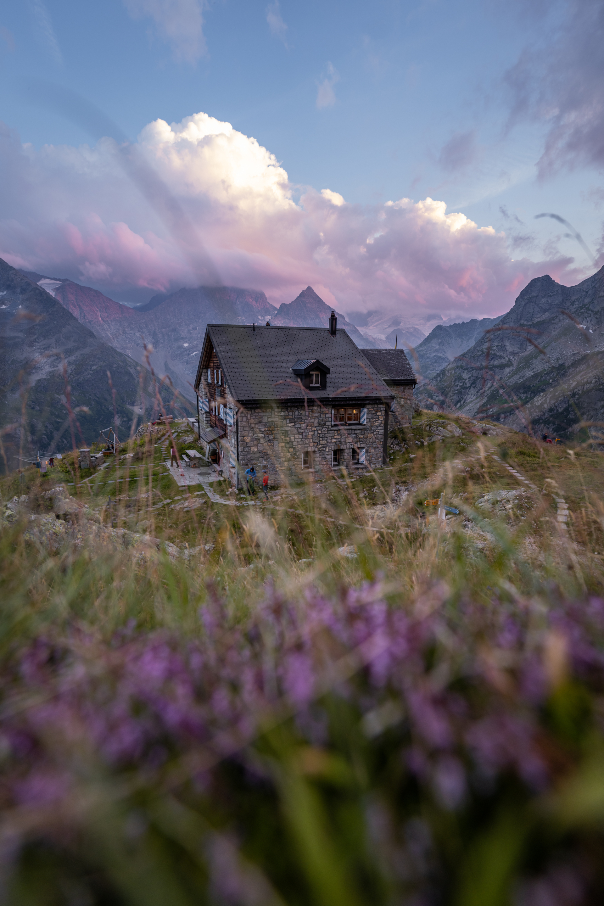 Ein verwittertes, altes Holzhaus steht einsam inmitten einer blühenden Wiese mit violetten Wildblumen. Im Hintergrund erheben sich eindrucksvolle Berggipfel, die von dramatischen Wolken und dem sanften Licht der Abenddämmerung umrahmt werden und der Szene eine ruhige, beinahe mystische Stimmung verleihen.