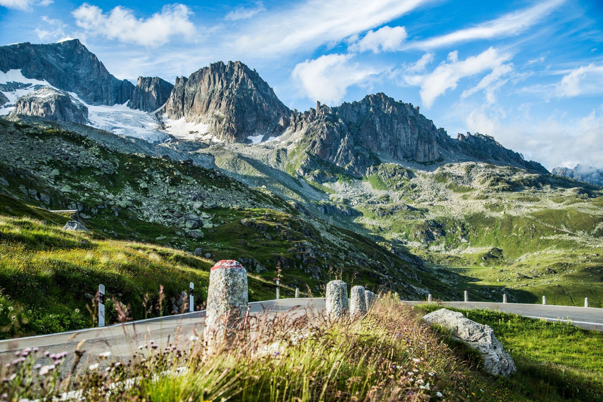 Eine kurvige Straße schlängelt sich durch die beeindruckende Berglandschaft des Furkapasses, umgeben von schroffen Gipfeln und saftigem Grün unter einem strahlend blauen Himmel. Die Szene vermittelt Abenteuer und die Schönheit der alpinen Natur.